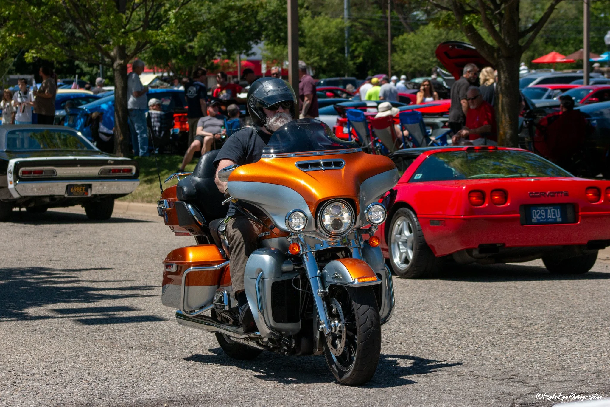 A man wearing a helmet and sunglasses riding an orange motorcycle in a crowded outdoor car show or event, with numerous parked cars, people, trees, and vendor tents in the background.