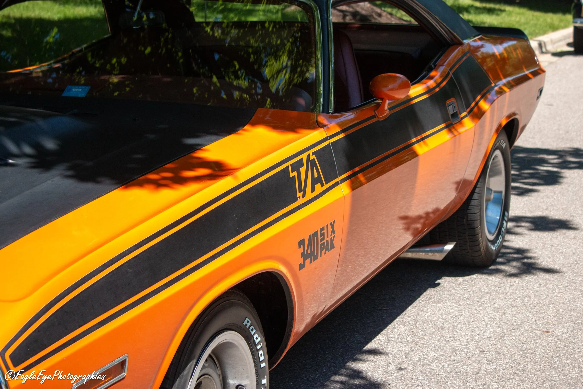 A vintage orange and black Dodge Charger muscle car parked outdoors on a sunny day, with green trees reflected on its windshield and hood.