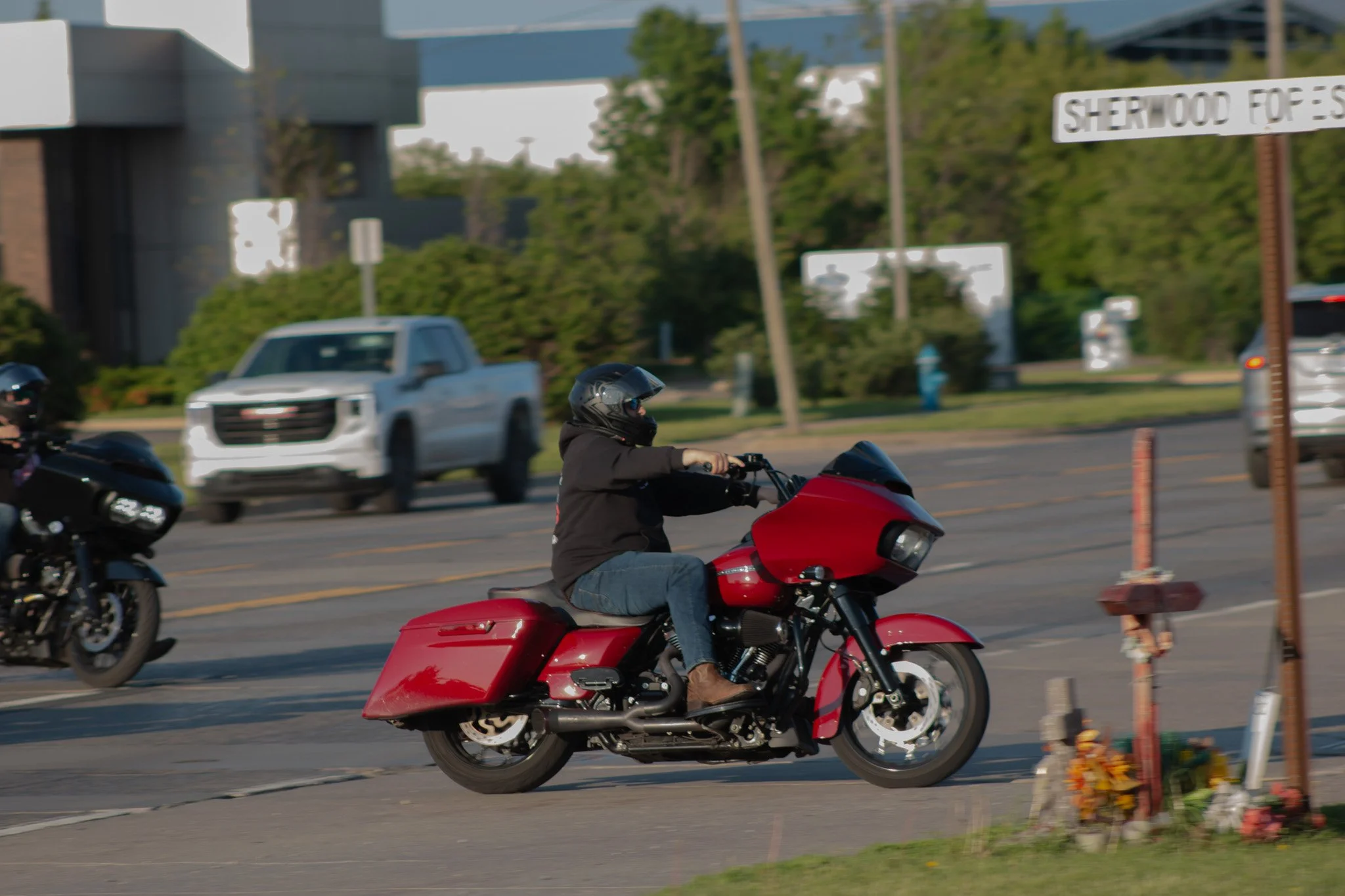 A person riding a red motorcycle on the street, wearing a black helmet and black jacket, with cars and a building in the background.