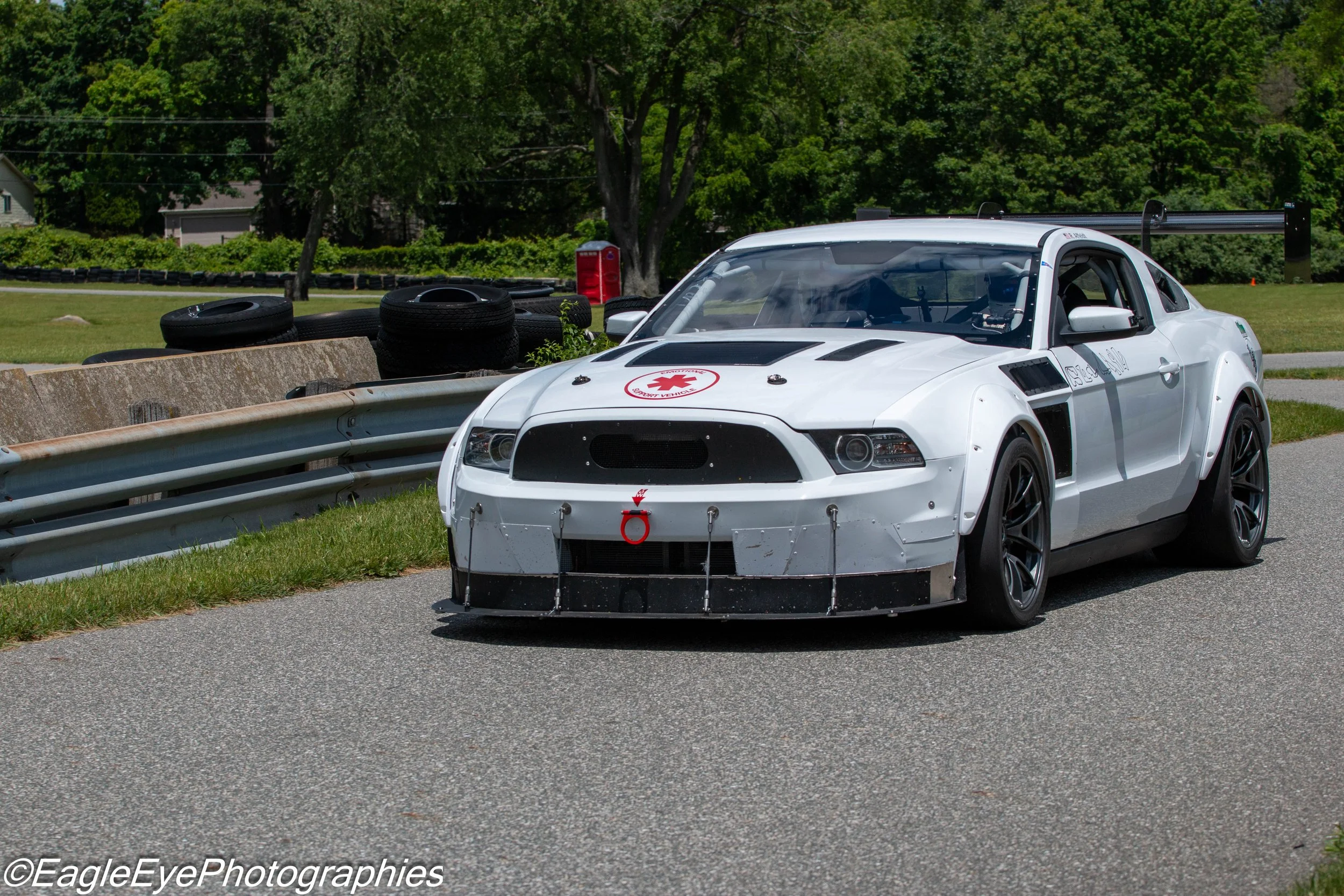 A white race car with racing decals and a large rear wing on a race track surrounded by green trees and tires as safety barriers.