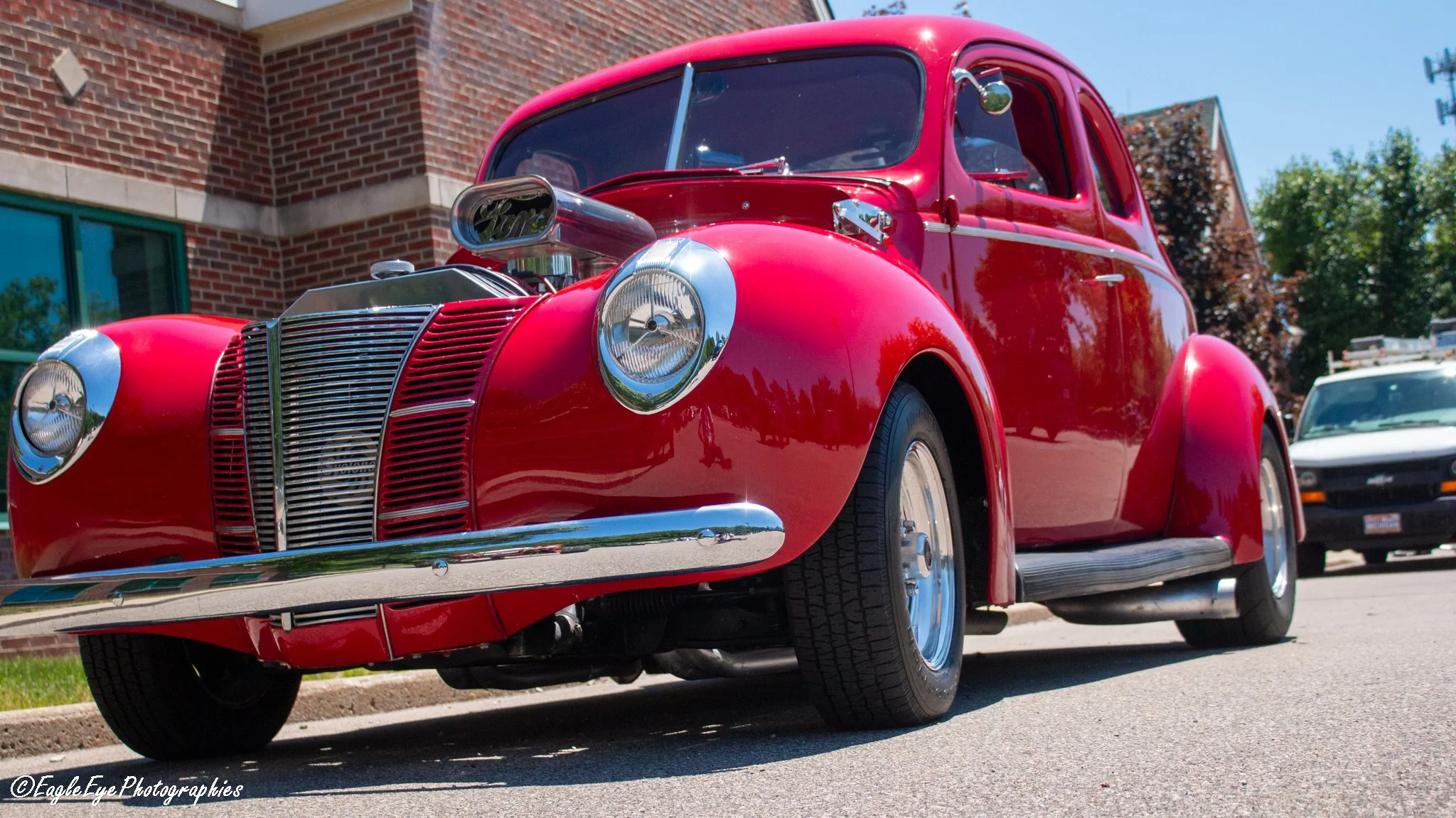A vintage red car with chrome details parked outdoors in front of a building with a brick wall, trees, and a clear blue sky.