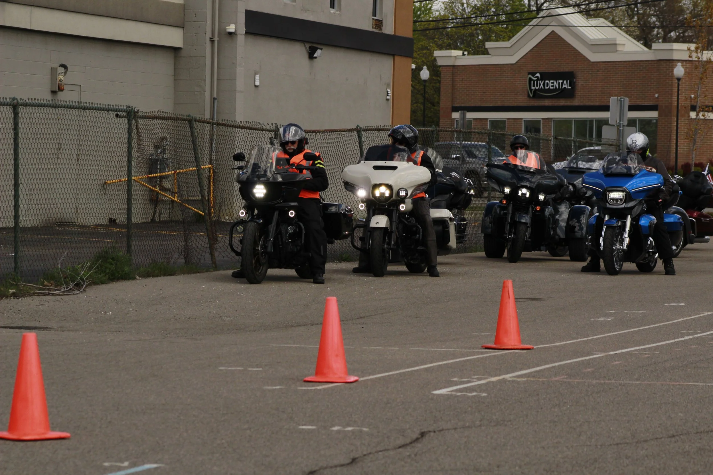 Group of motorcyclists wearing helmets and reflective vests, lined up behind orange traffic cones in an outdoor lot.