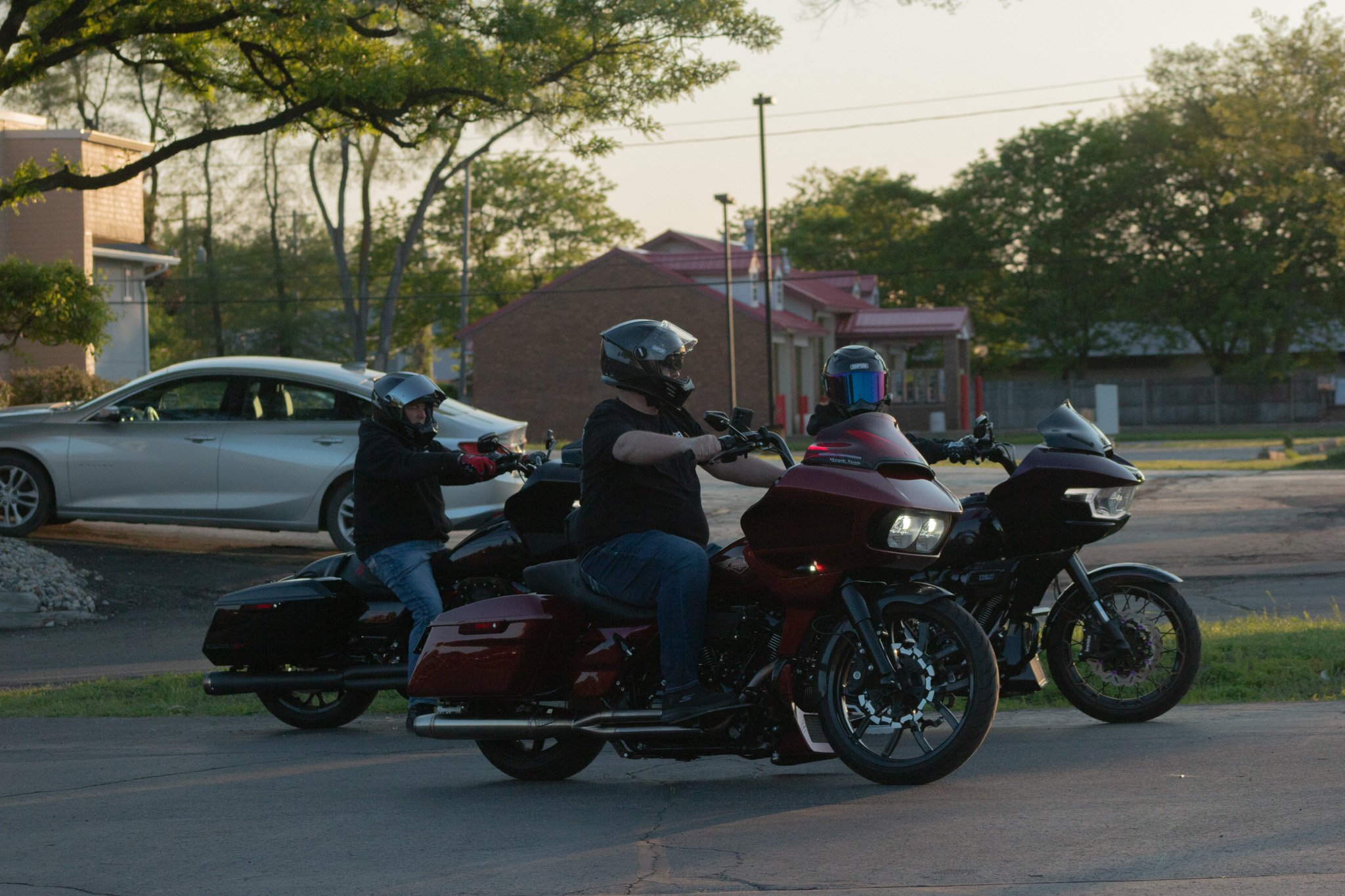 Three motorcyclists wearing helmets riding on a road during sunset with a background of trees, houses, and a car.