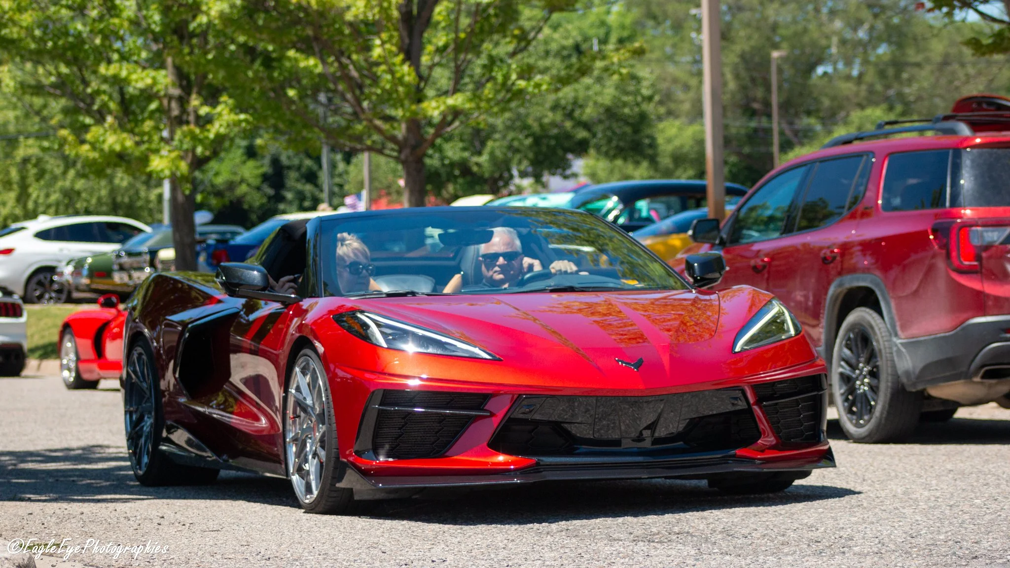 A red sports car driving through a parking lot with trees in the background and other parked vehicles nearby.
