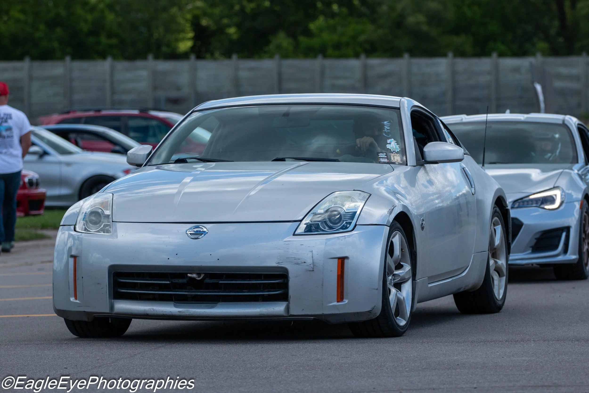 A silver Nissan sports car parked in a parking lot with other cars and a wooden fence in the background.