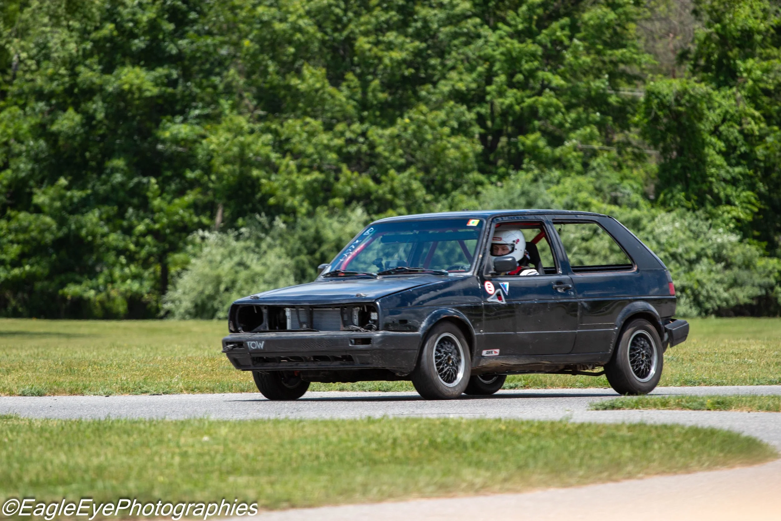 A person in a race car reaching a turn on a grassy racetrack, with trees in the background.
