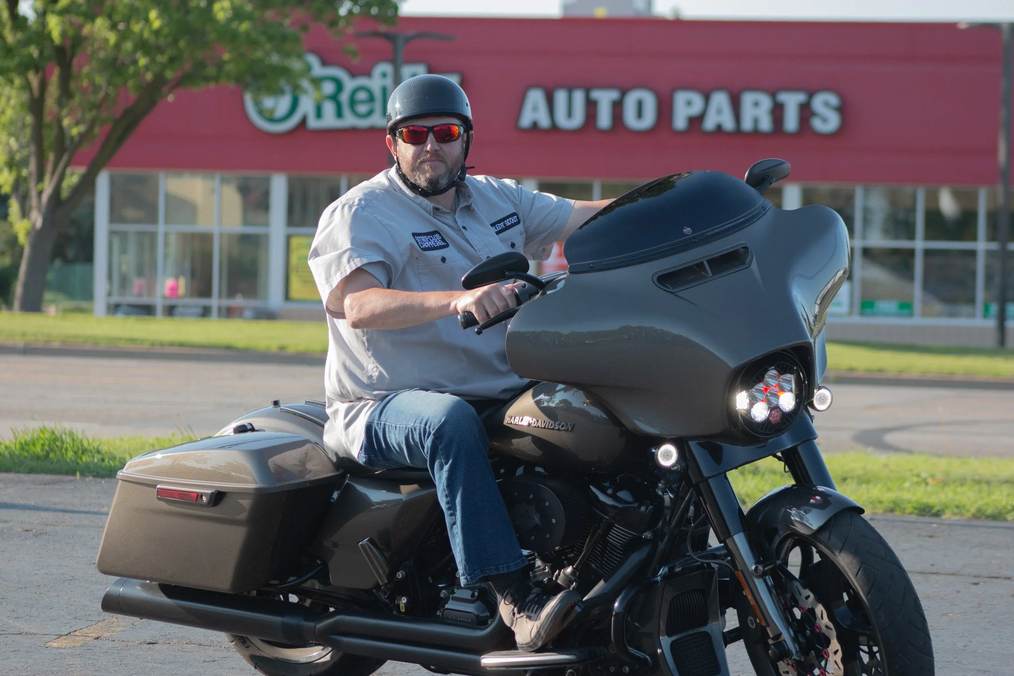 A man wearing a black helmet, sunglasses, and a beige shirt with patches, riding a black Harley Davidson motorcycle outside near an auto parts store with a red sign and green trees.