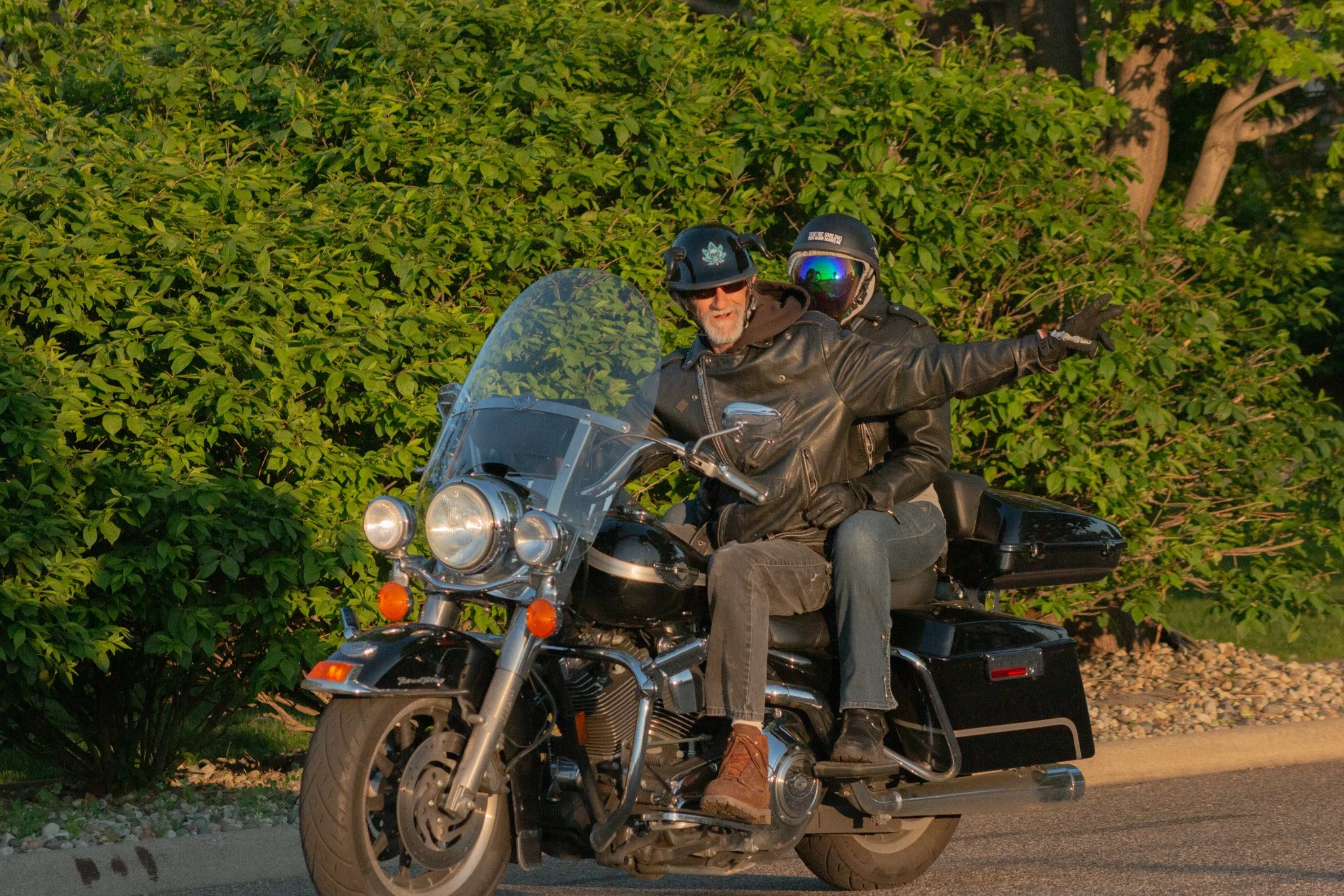Two men riding a motorcycle with a sidecar, wearing helmets and leather jackets, near lush green bushes and trees.