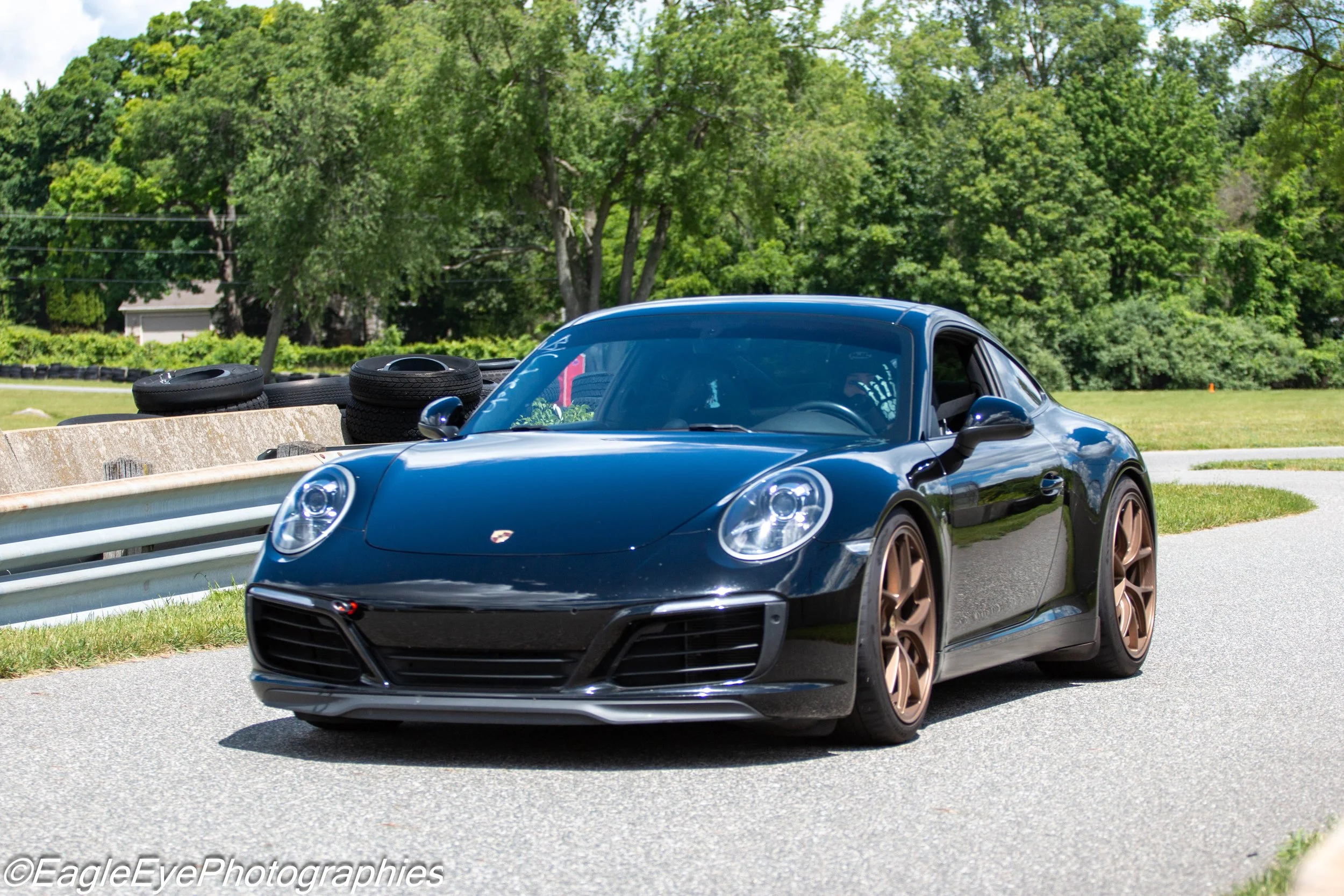 A black sports car with bronze wheels on the side of a road, with trees and green foliage in the background.