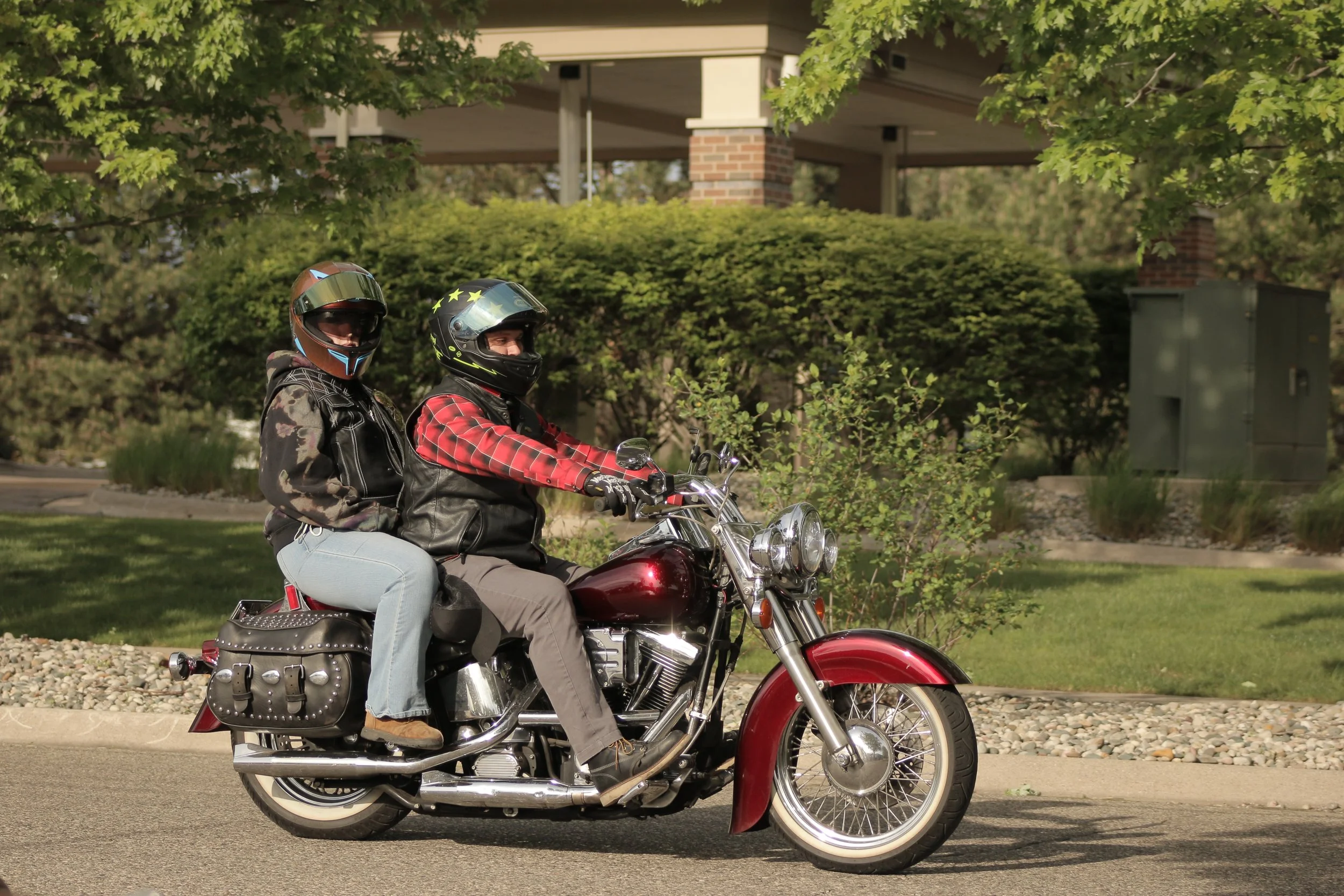 Two women wearing helmets ride a red motorcycle on a suburban street with greenery and a house in the background.
