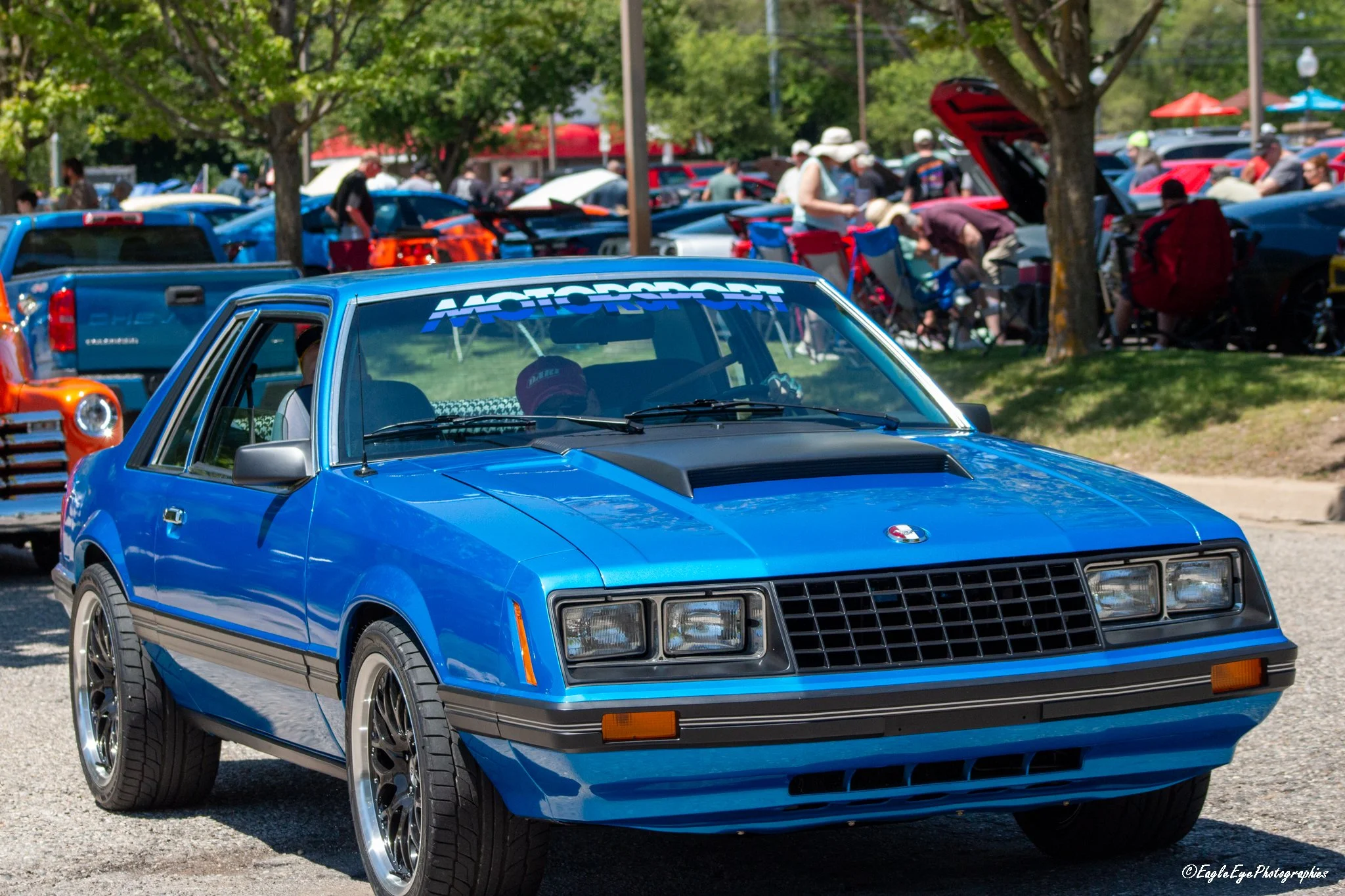 A bright blue vintage sports car with a black hood parked at a car show, with people and other vehicles visible in the background under trees and sunny weather.