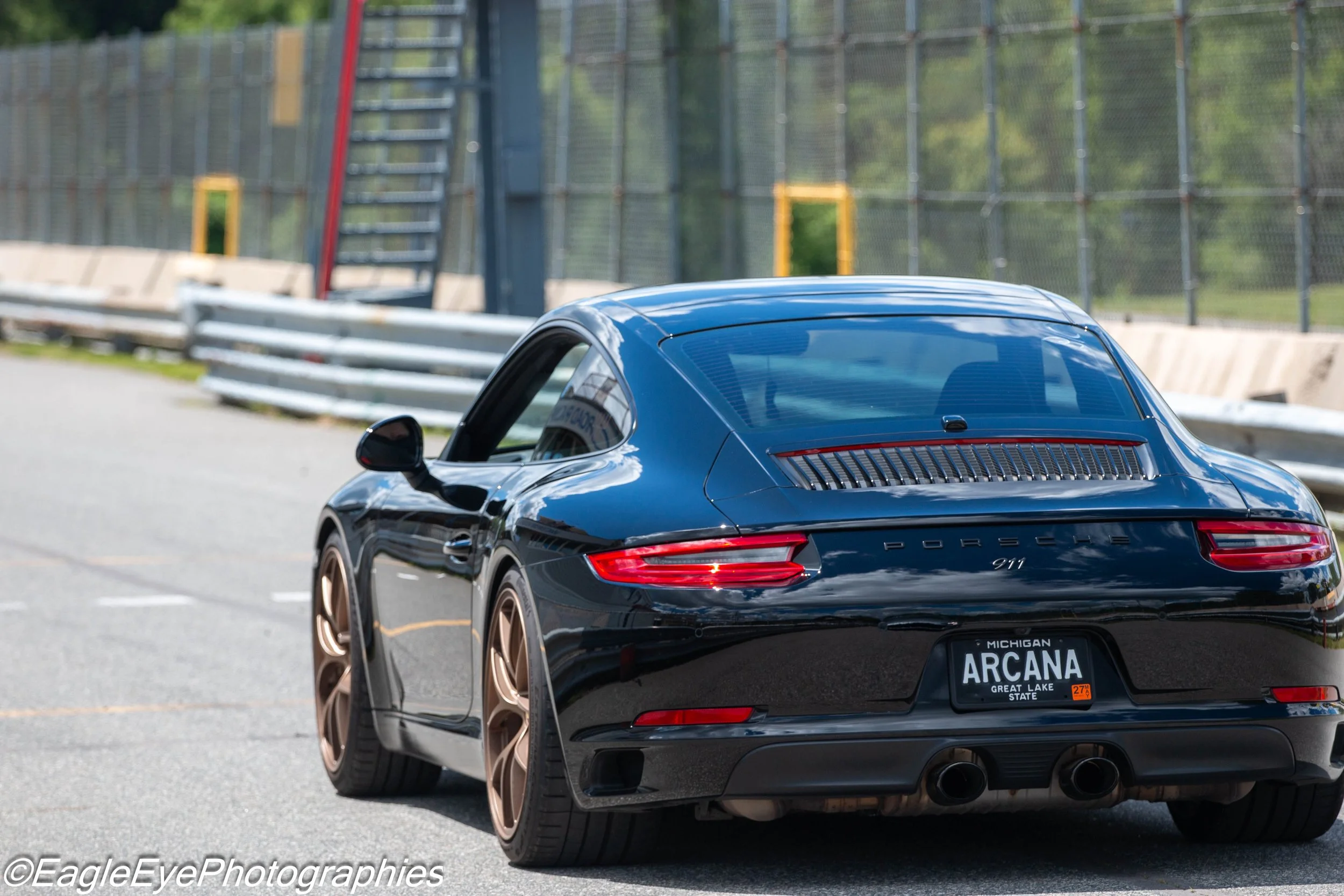Black Porsche 911 with Michigan license plate reading 'Arcona' parked on a paved area near a fence with greenery in the background.