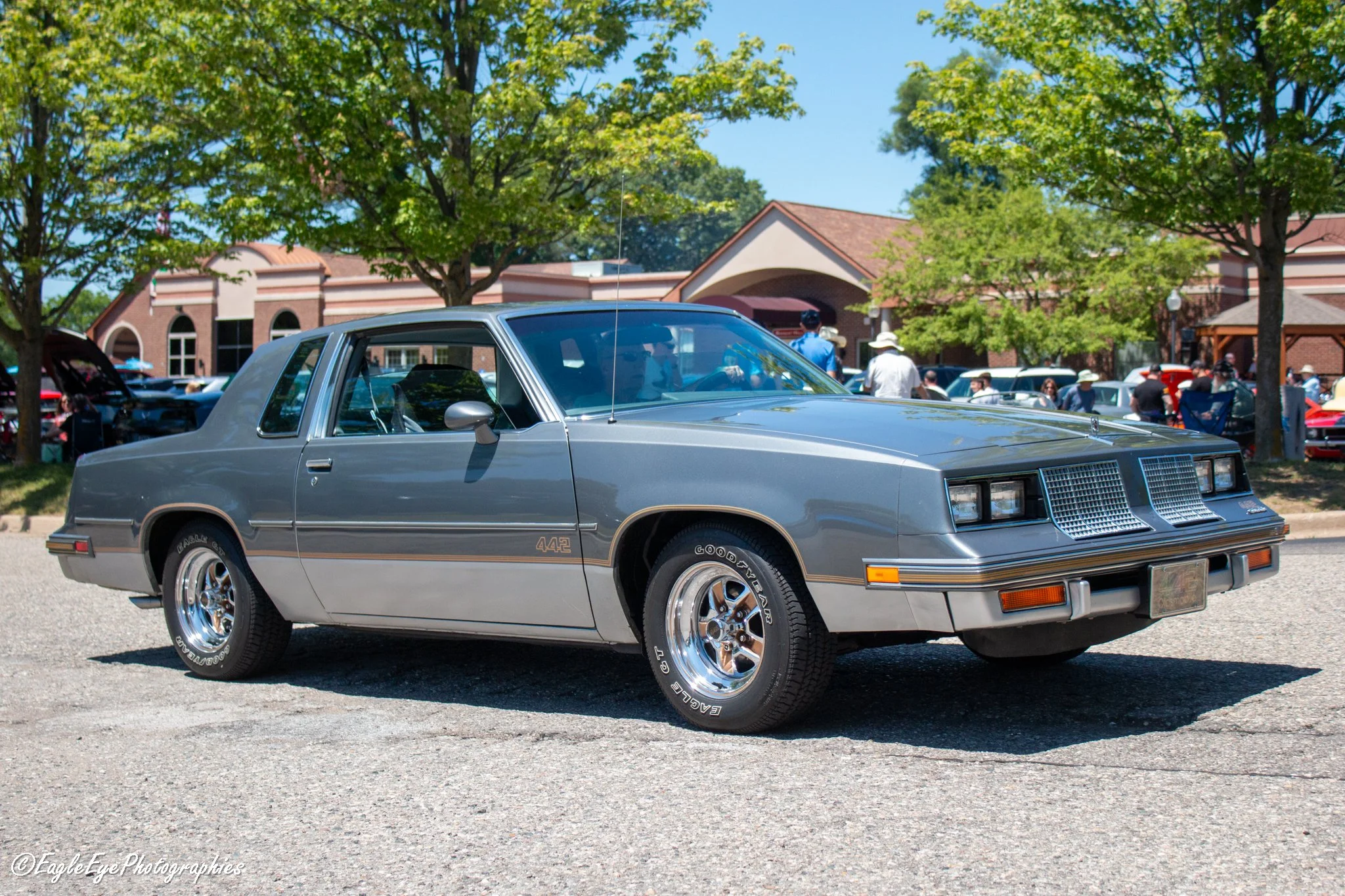 A silver vintage car parked outdoors at a car show with trees, a building, and people in the background on a sunny day.