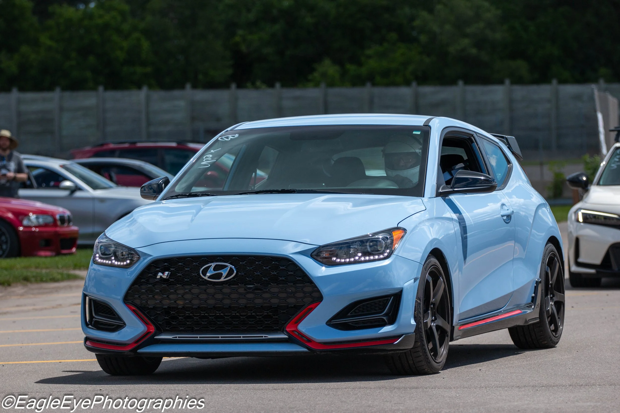 A blue Hyundai Veloster N sports car parked outdoors during the daytime with a black grille, LED headlights, and red accents on the front bumper. Several other vehicles and people are visible in the background, with a wooden fence and green trees beh