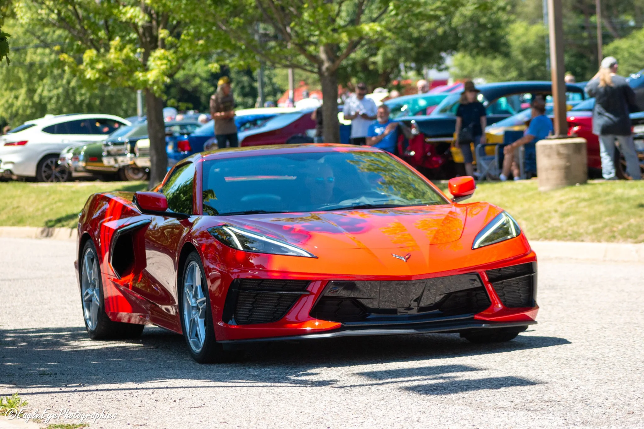 A shiny red sports car, likely a Chevrolet Corvette, parked on a street during a car show under trees. In the background, there are people walking and looking at other cars and a lot of parked vehicles.