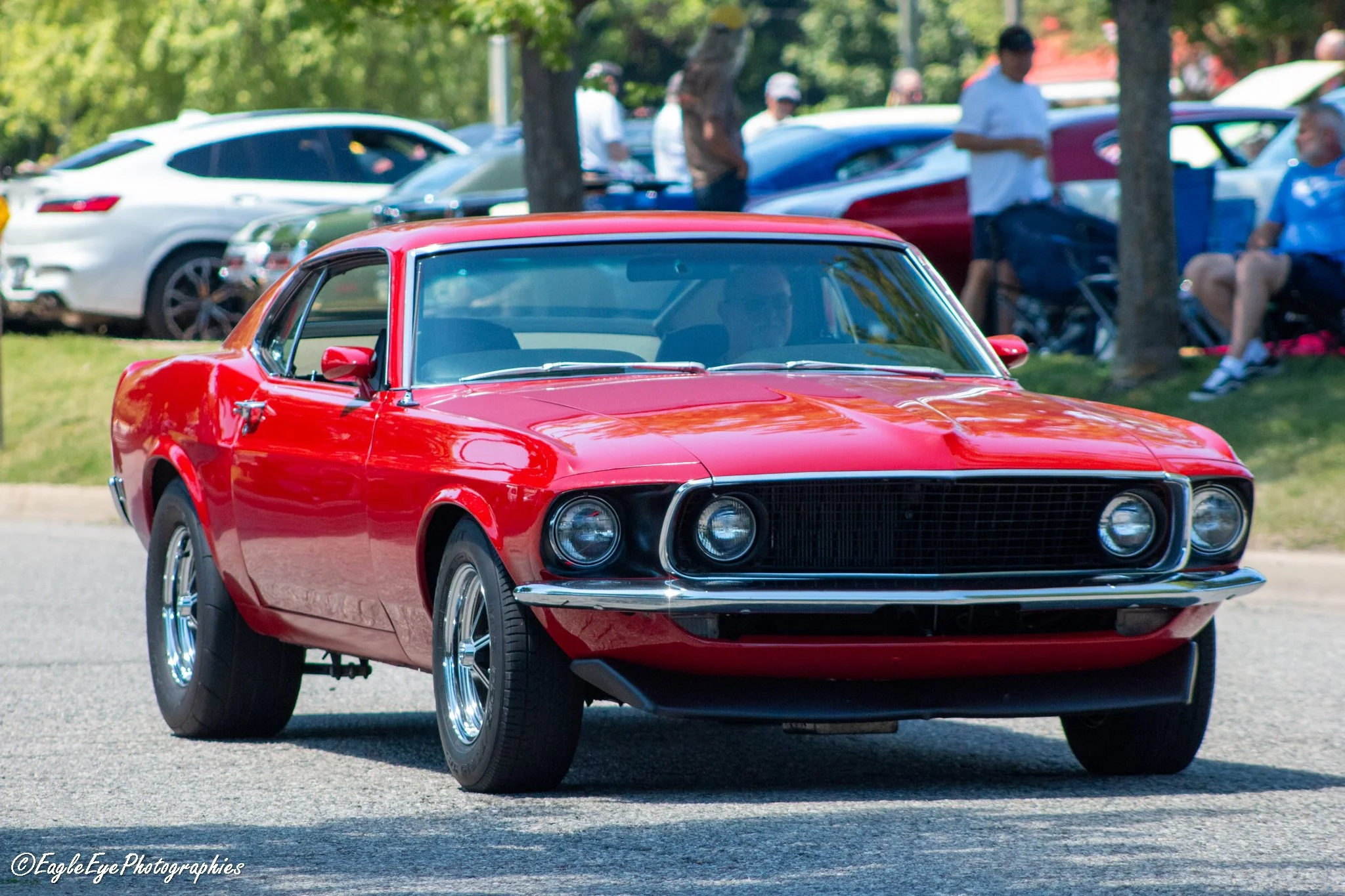 A red vintage car driving on a street with a group of people and other cars in the background, some sitting under trees and standing.