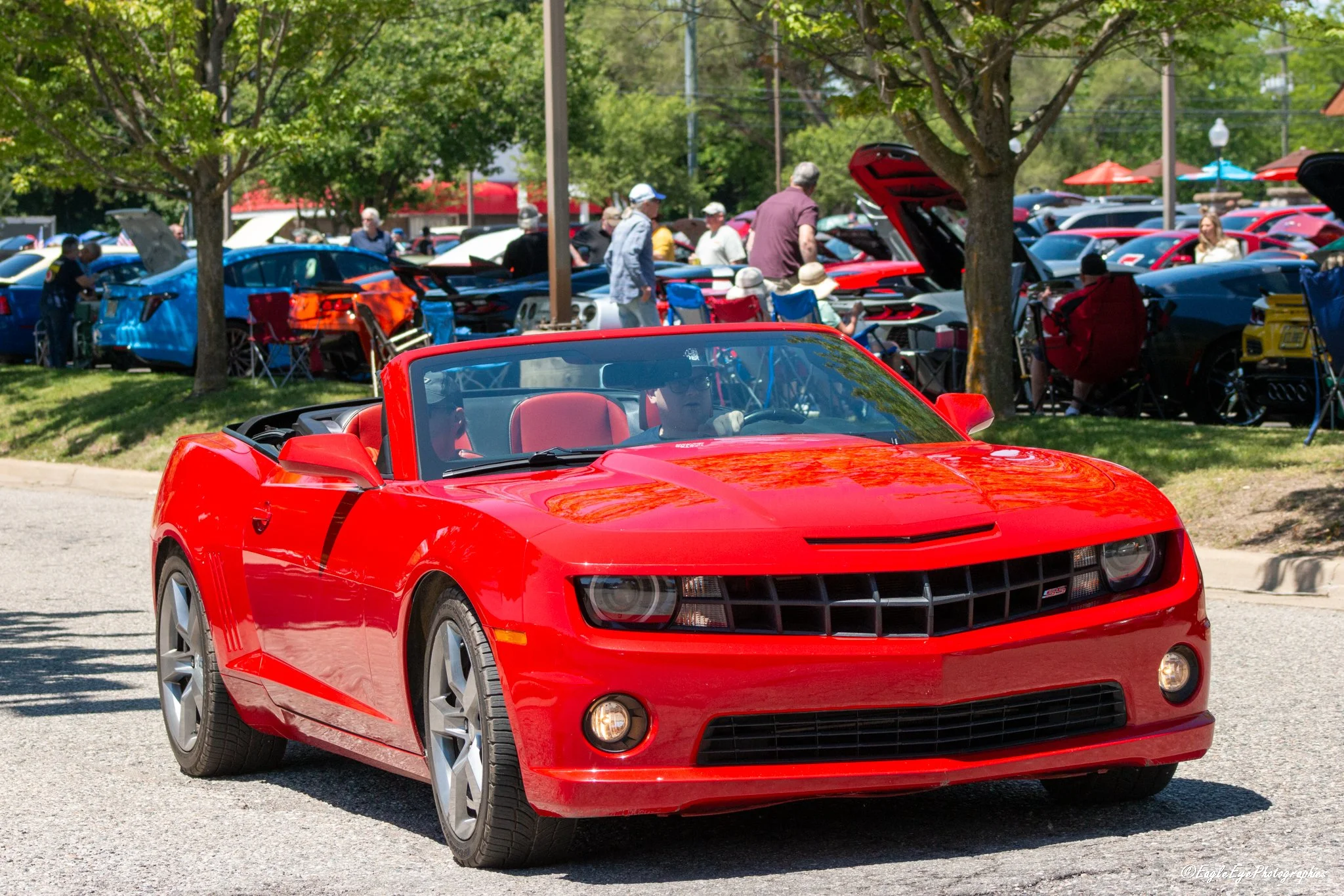 A bright red Chevrolet Camaro convertible driving past a classic car show with various cars and people in the background.