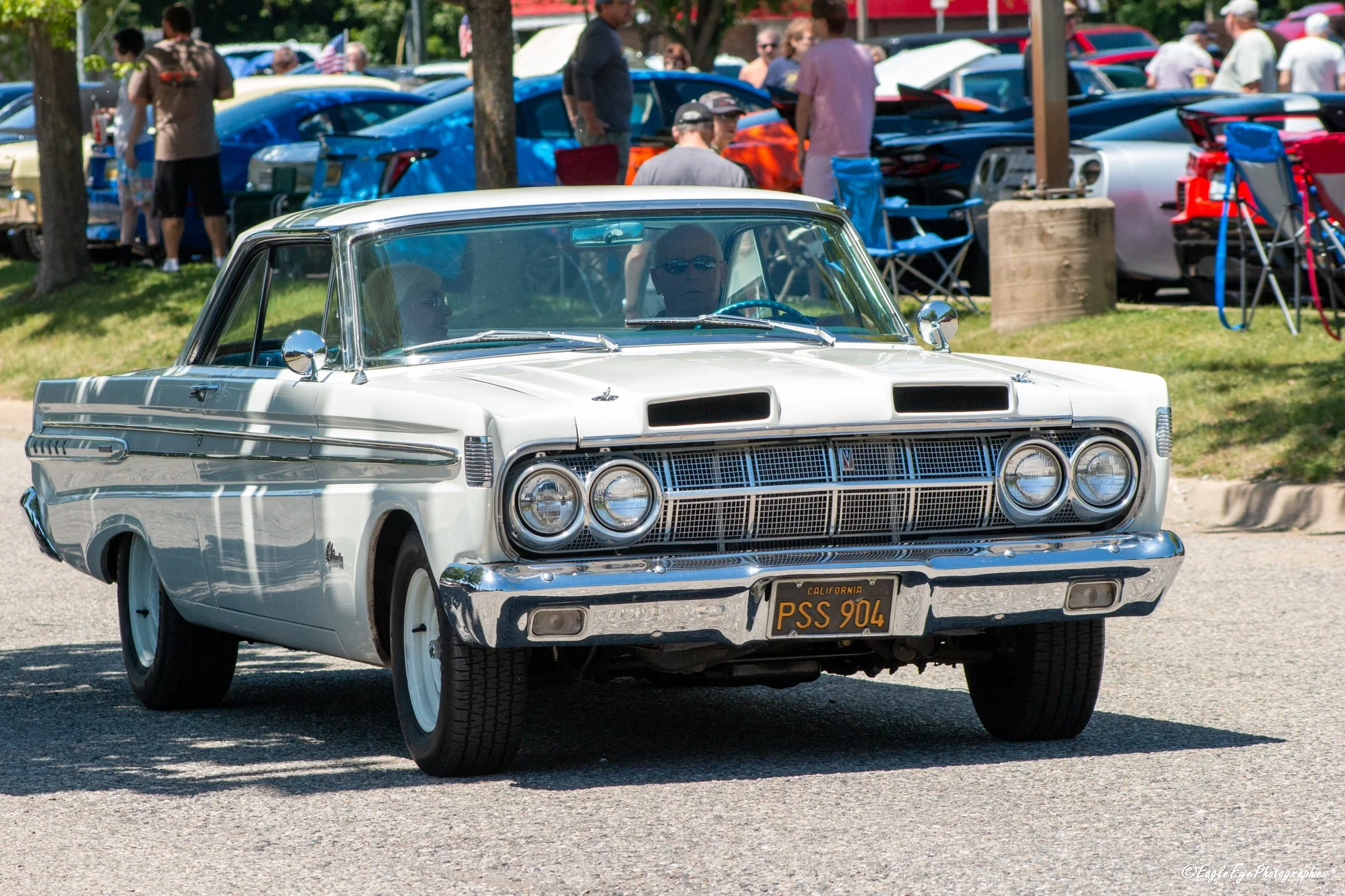 A vintage white car with a California license plate driving on a street during a car show or parade, with people and other cars in the background.