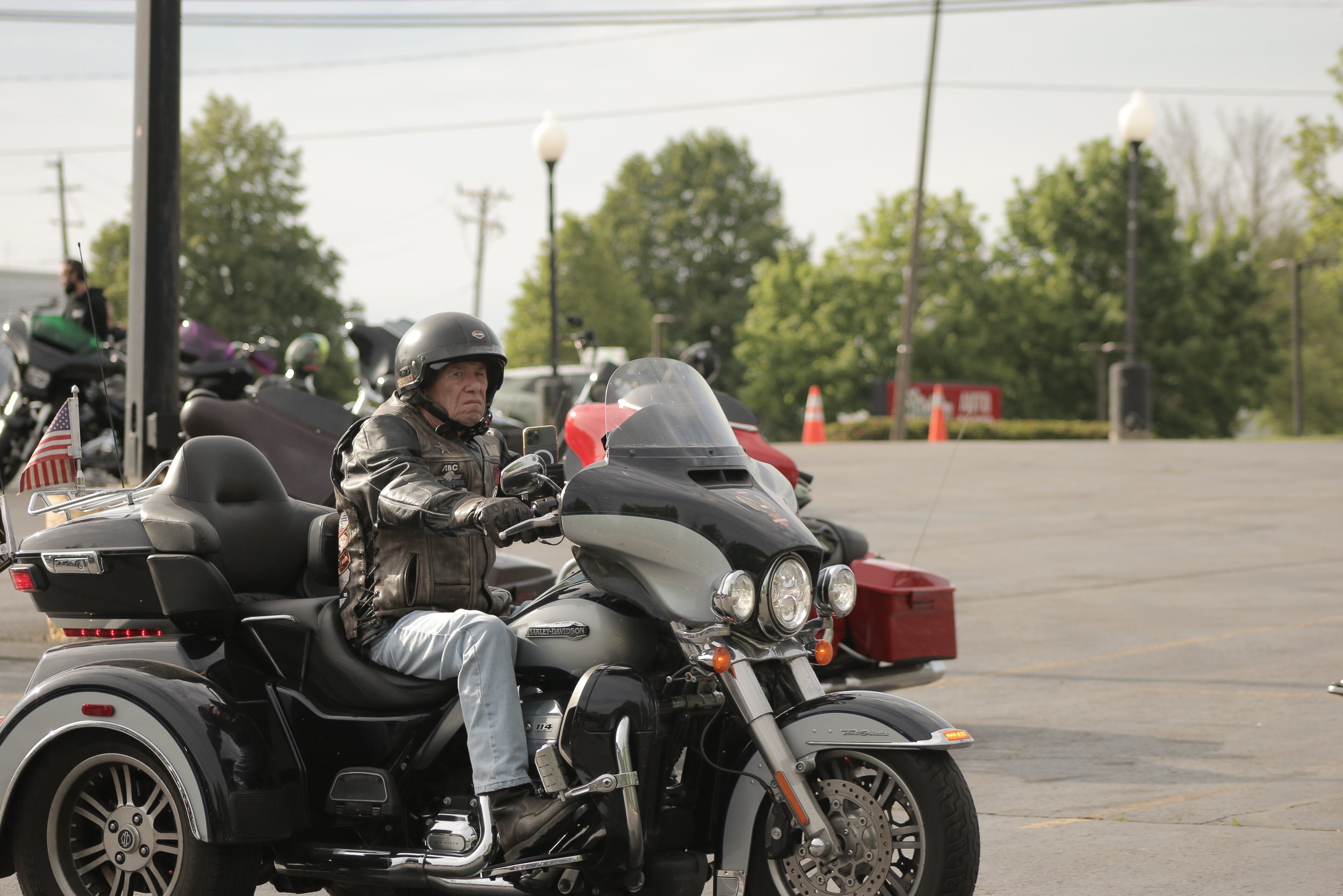 A man wearing a black helmet and leather jacket riding a black Harley Davidson trike motorcycle with an American flag on the back. He is holding a phone in his right hand. The background shows other motorcycles, trees, and traffic cones in a parking 