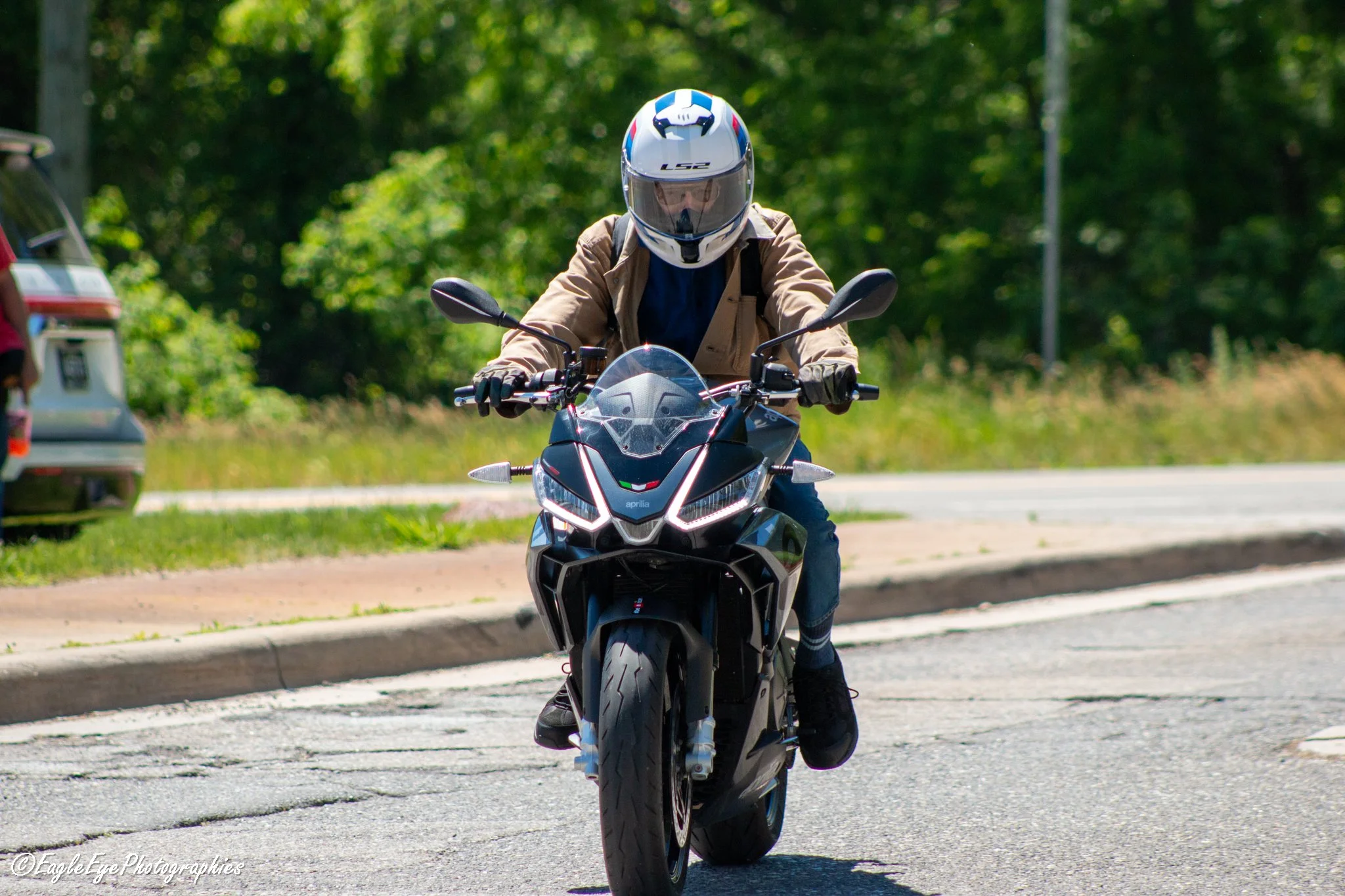 A person wearing a full-face helmet and brown jacket riding a black motorcycle on a paved road with green trees in the background.