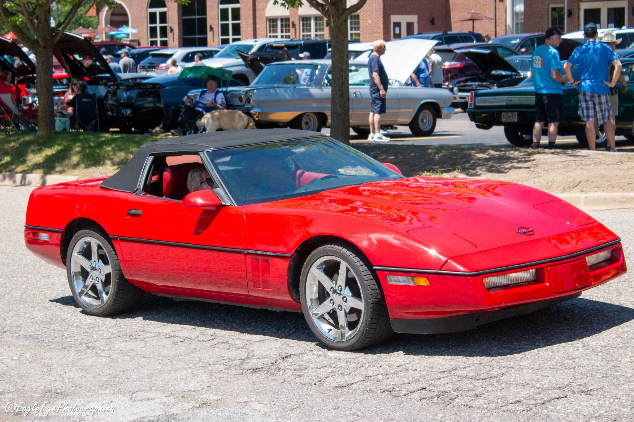 A red Chevrolet Corvette with a black top parked outdoors at a car show. In the background, several classic cars and a gathering of people are visible, some looking at cars with hoods open, others sitting or talking near trees and parked cars.