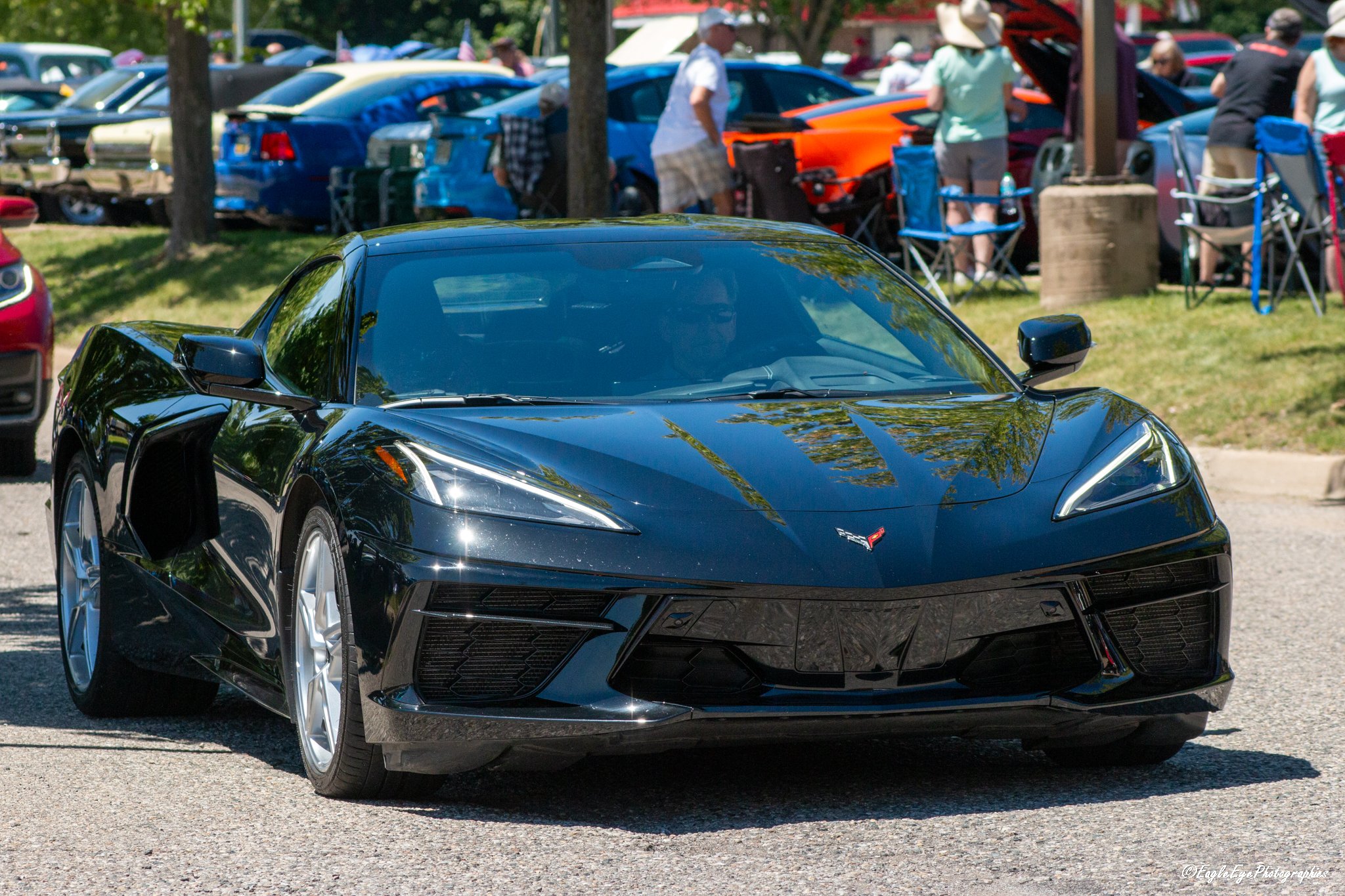 A black sports car parked on the street during an outdoor car show or gathering. People are seen in the background near parked cars and tents on a grassy area, some looking under the hood of a car, with trees and flags visible.