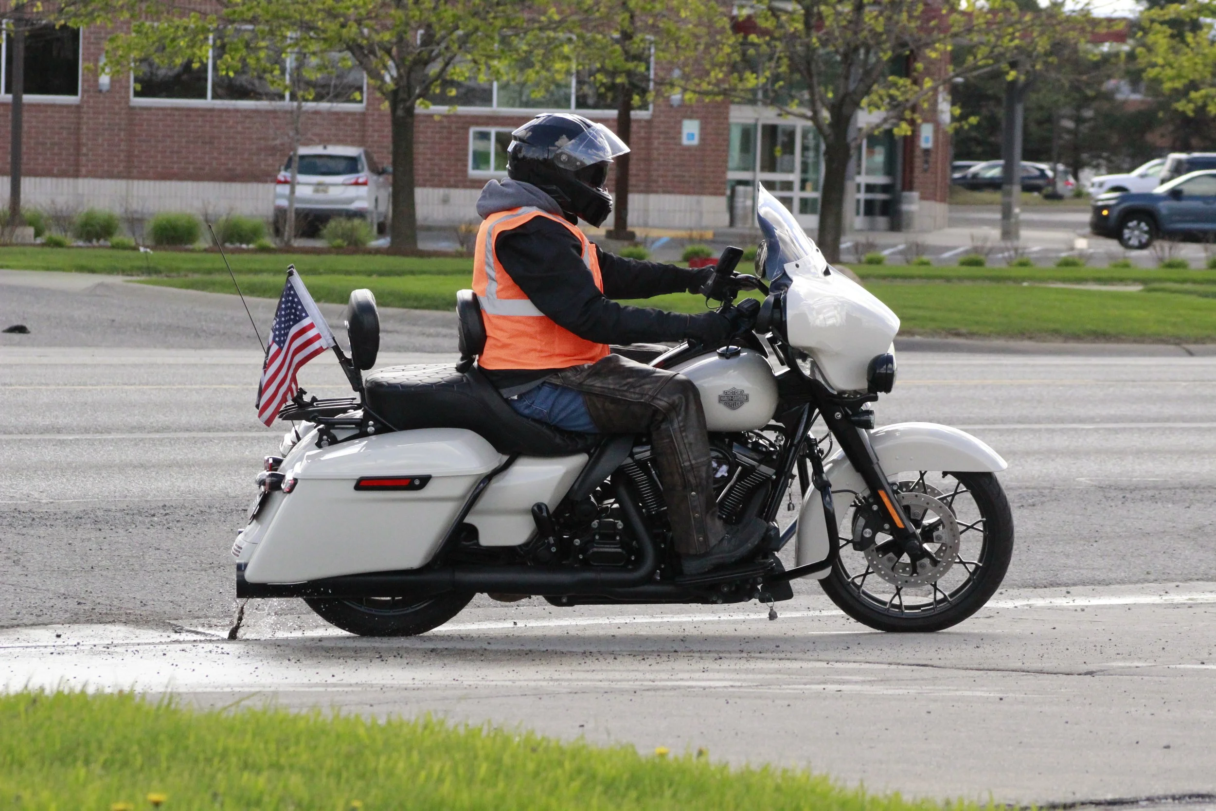 A person riding a white Harley Davidson motorcycle on a city street, wearing a black helmet, orange safety vest, and jeans, with a small American flag attached to the back of the motorcycle.