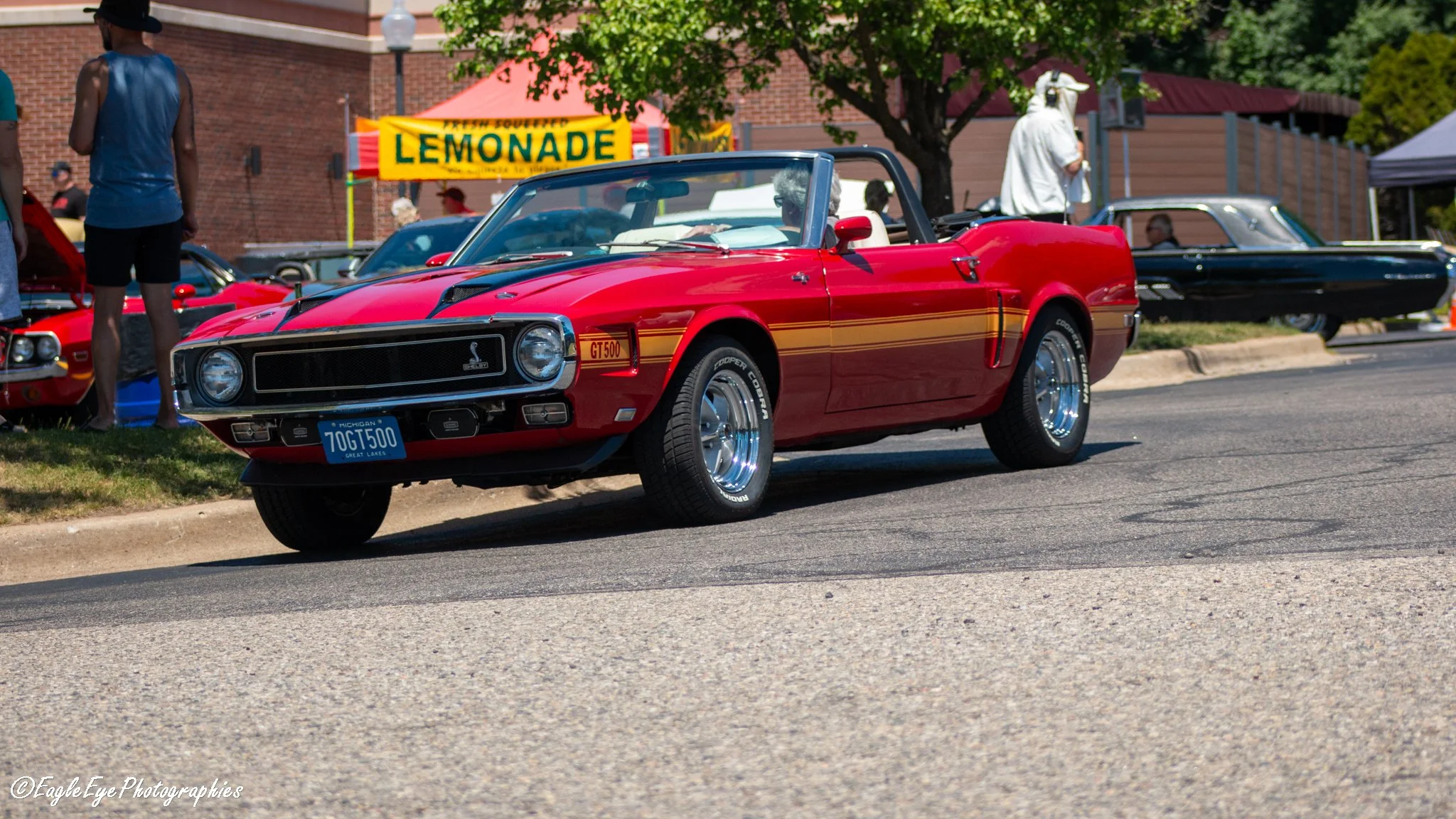 Red convertible sports car with a Michigan license plate, parked on the street at a car show, with people and vintage cars in the background.