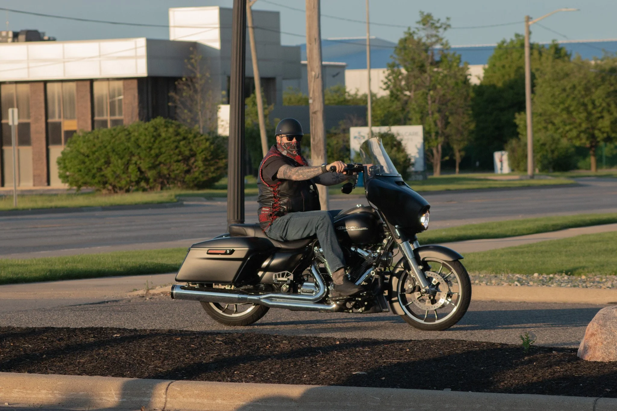 A man riding a black motorcycle on a street with buildings, trees, utility poles, and power lines in the background during daytime.