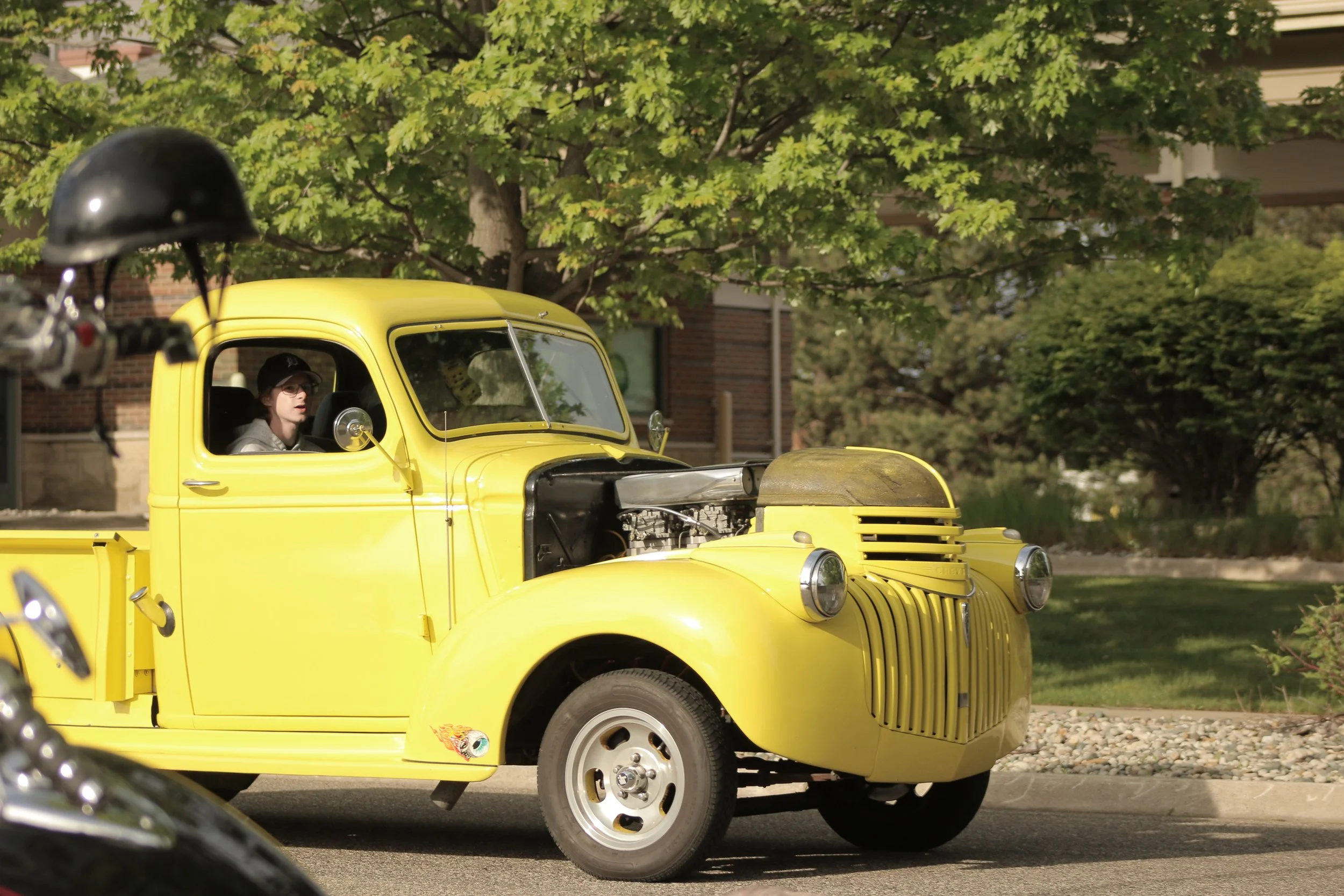 A person inside a bright yellow vintage truck, with the hood and engine visible, parked outdoors under green trees.