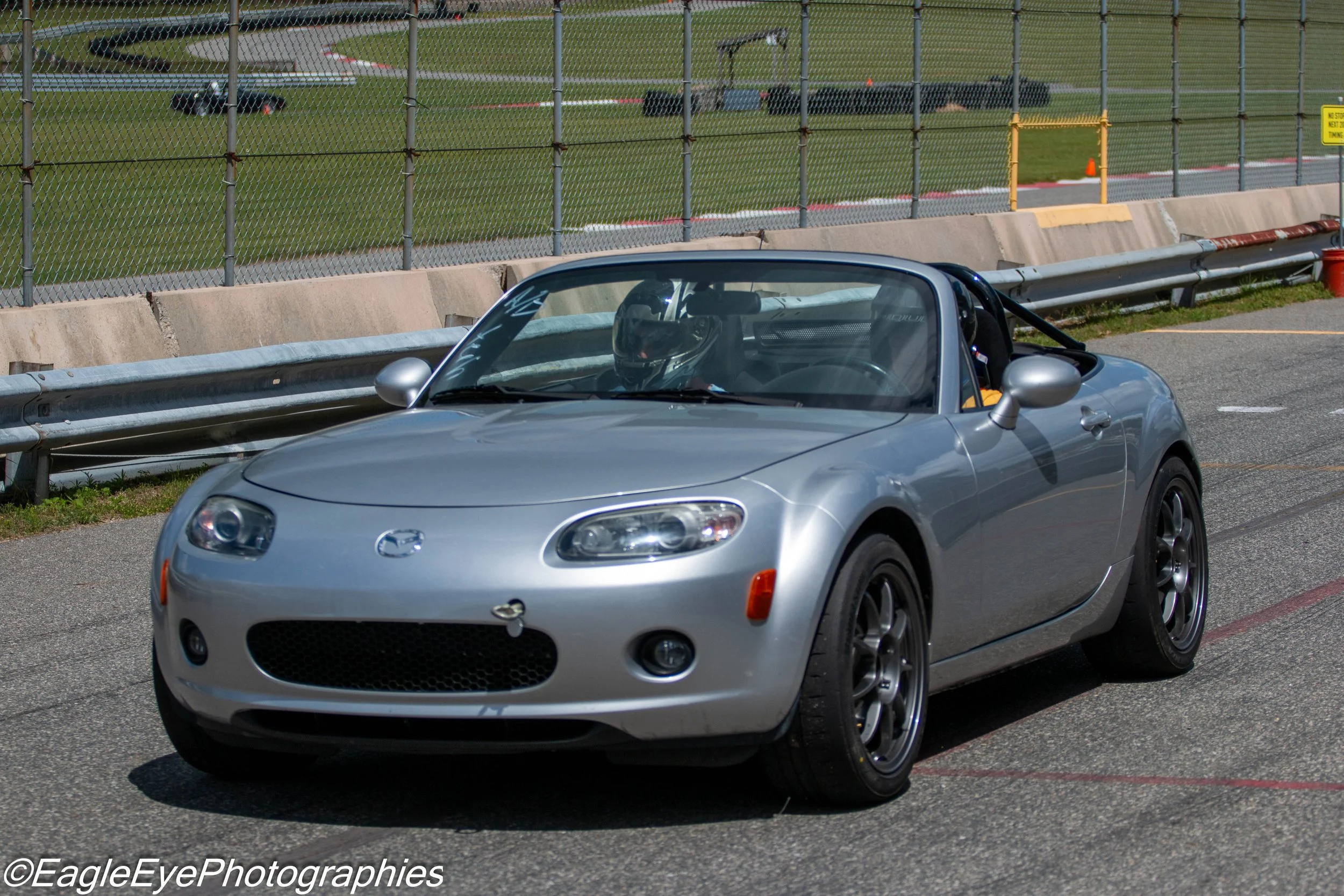 A silver Mazda MX-5 Miata sports car on a race track with a driver wearing a helmet, and a race track with a fence and grass in the background.