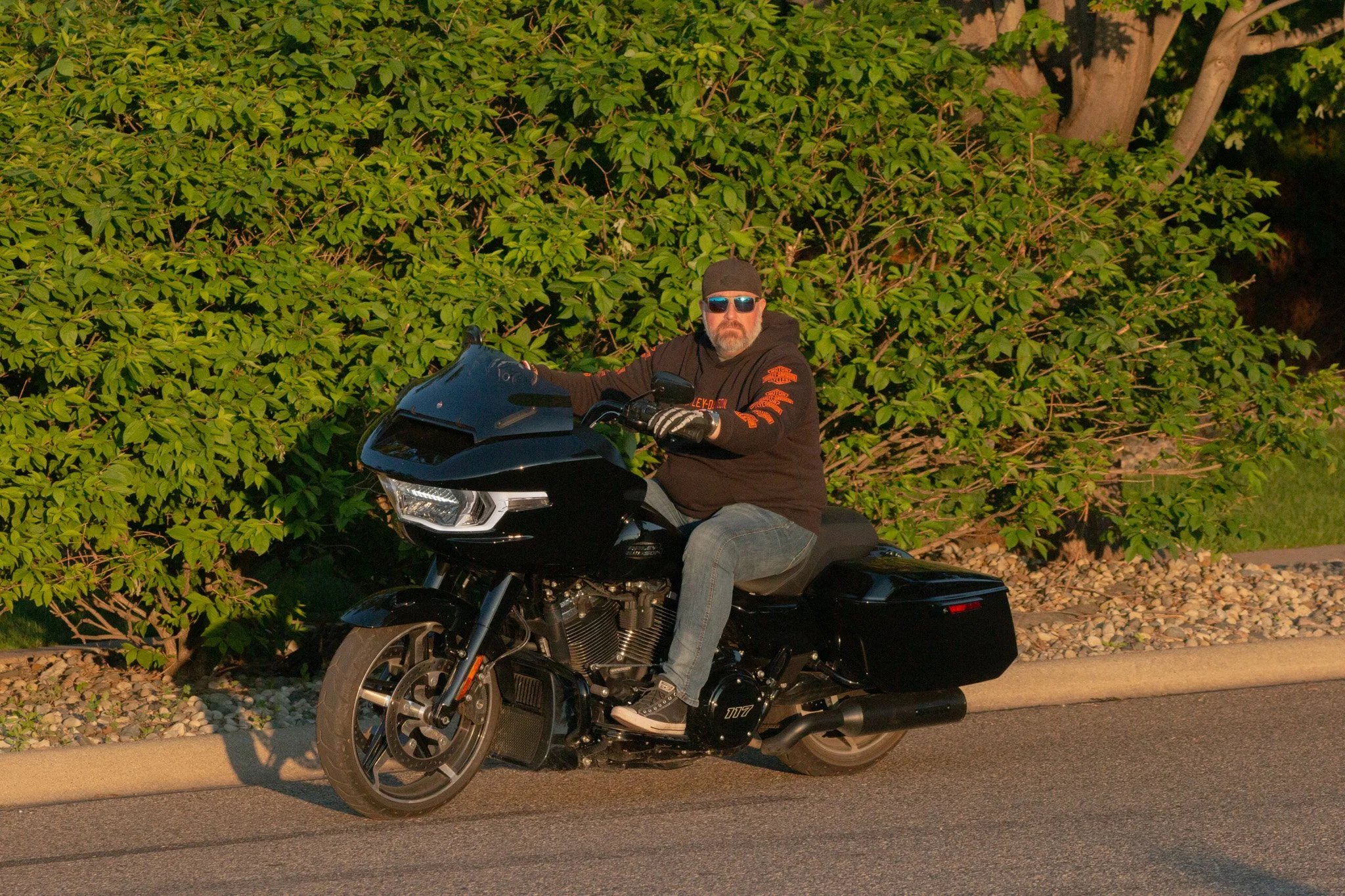 A man wearing sunglasses, a black hoodie with orange text, and a black cap is riding a black Harley-Davidson motorcycle along a roadway. He is positioned in front of a lush green bush with a large tree in the background. The sunlight suggests it is e