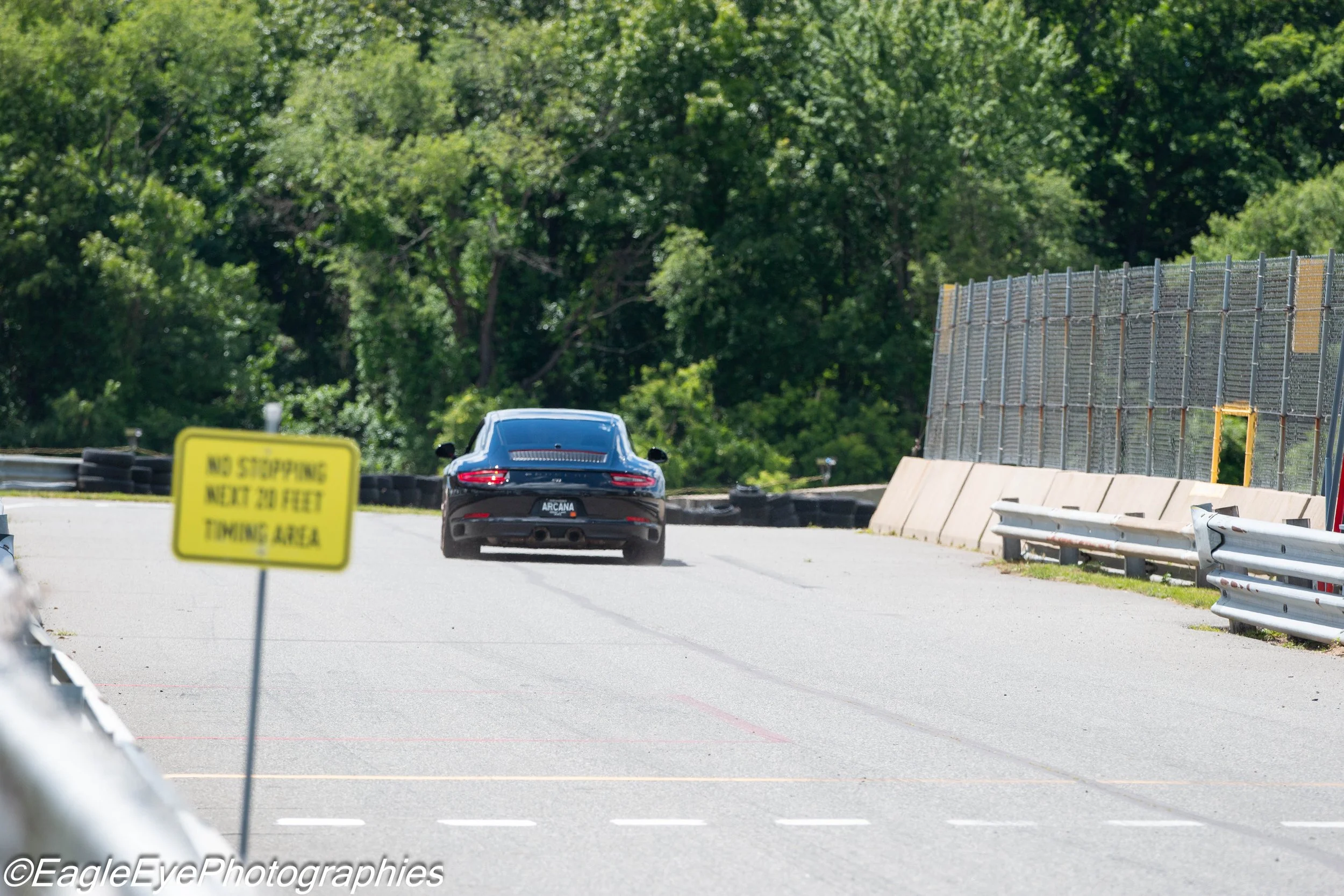 A black sports car driving on a racetrack near a safety barrier with a yellow sign that says 'No stopping next 20 feet towing area', surrounded by dense green trees.