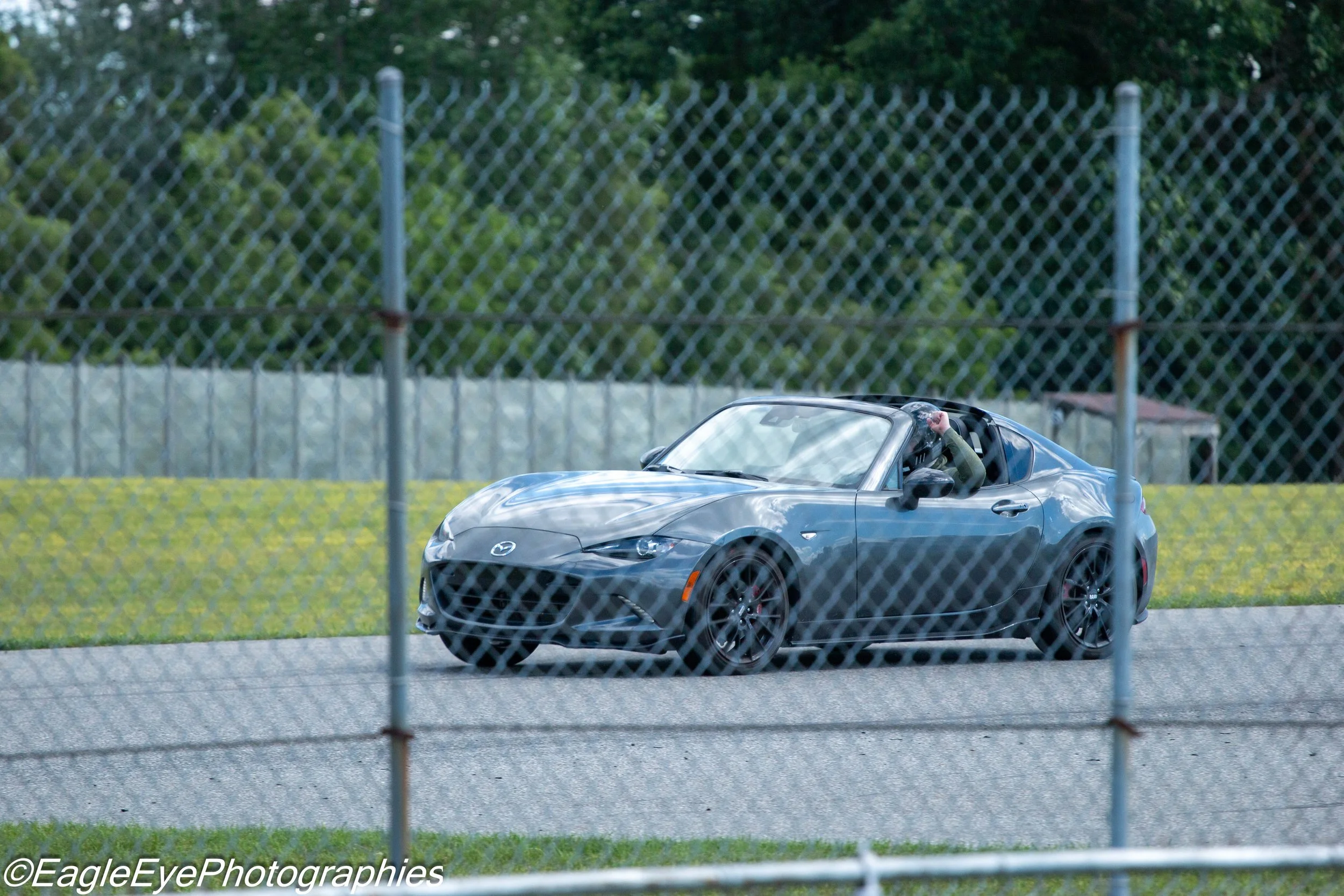 A blue Mazda MX-5 sports car on a road with greenery and a chain-link fence in the foreground.