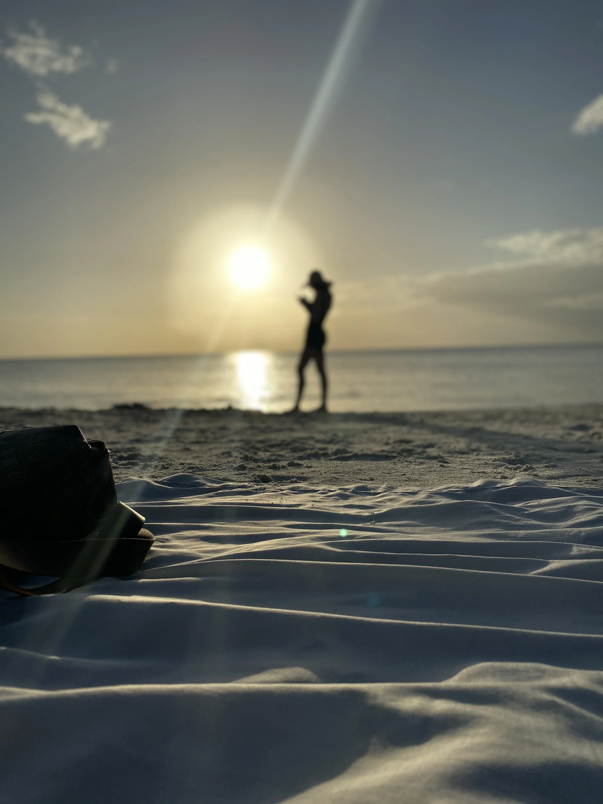 A person standing on a sandy beach, silhouetted against a setting sun, with the ocean in the background and a pair of shoes in the foreground.