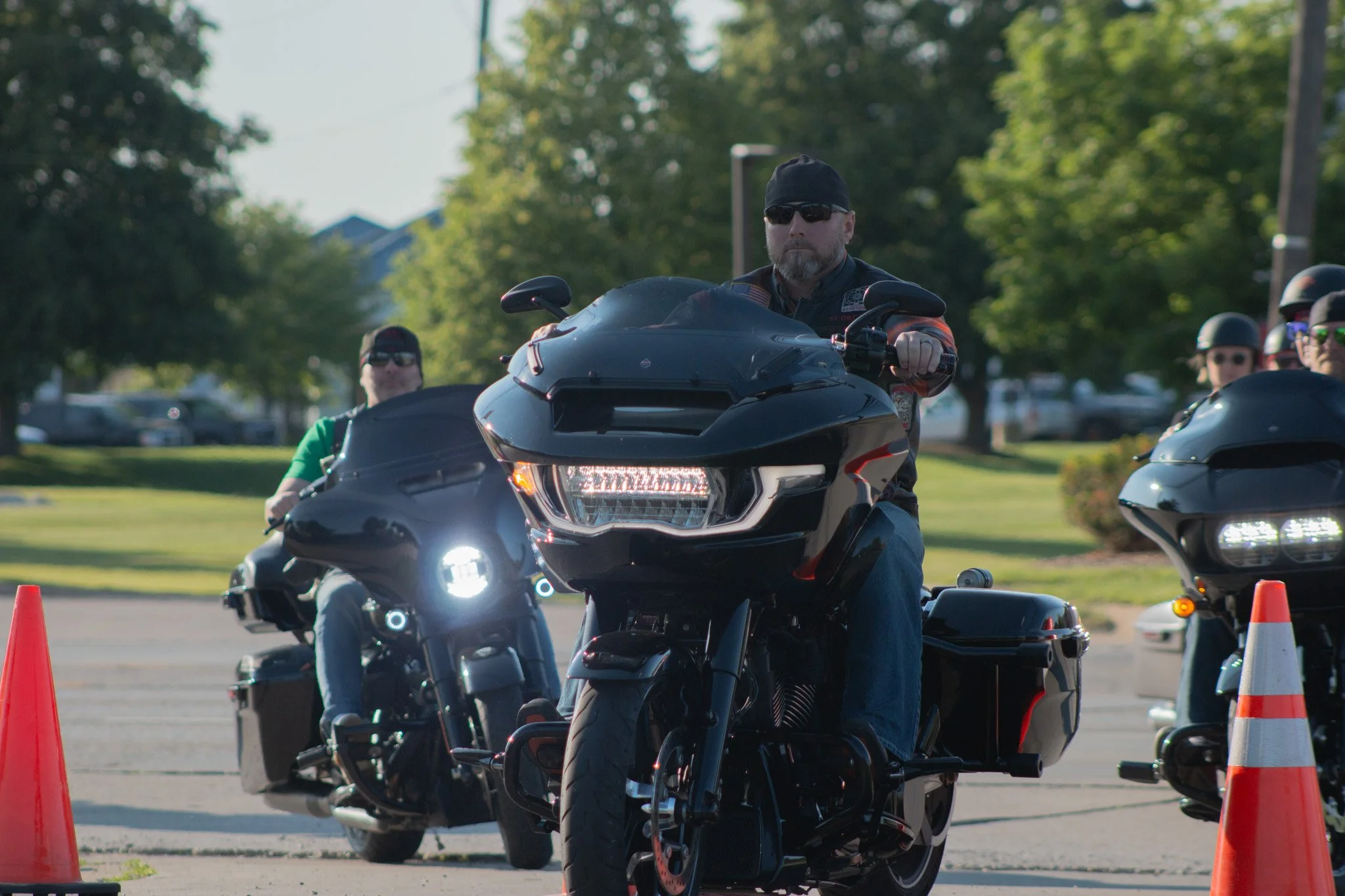 Man with beard wearing sunglasses and black cap riding a black motorcycle during a motorcycle safety event, with other riders and orange traffic cones in the background.