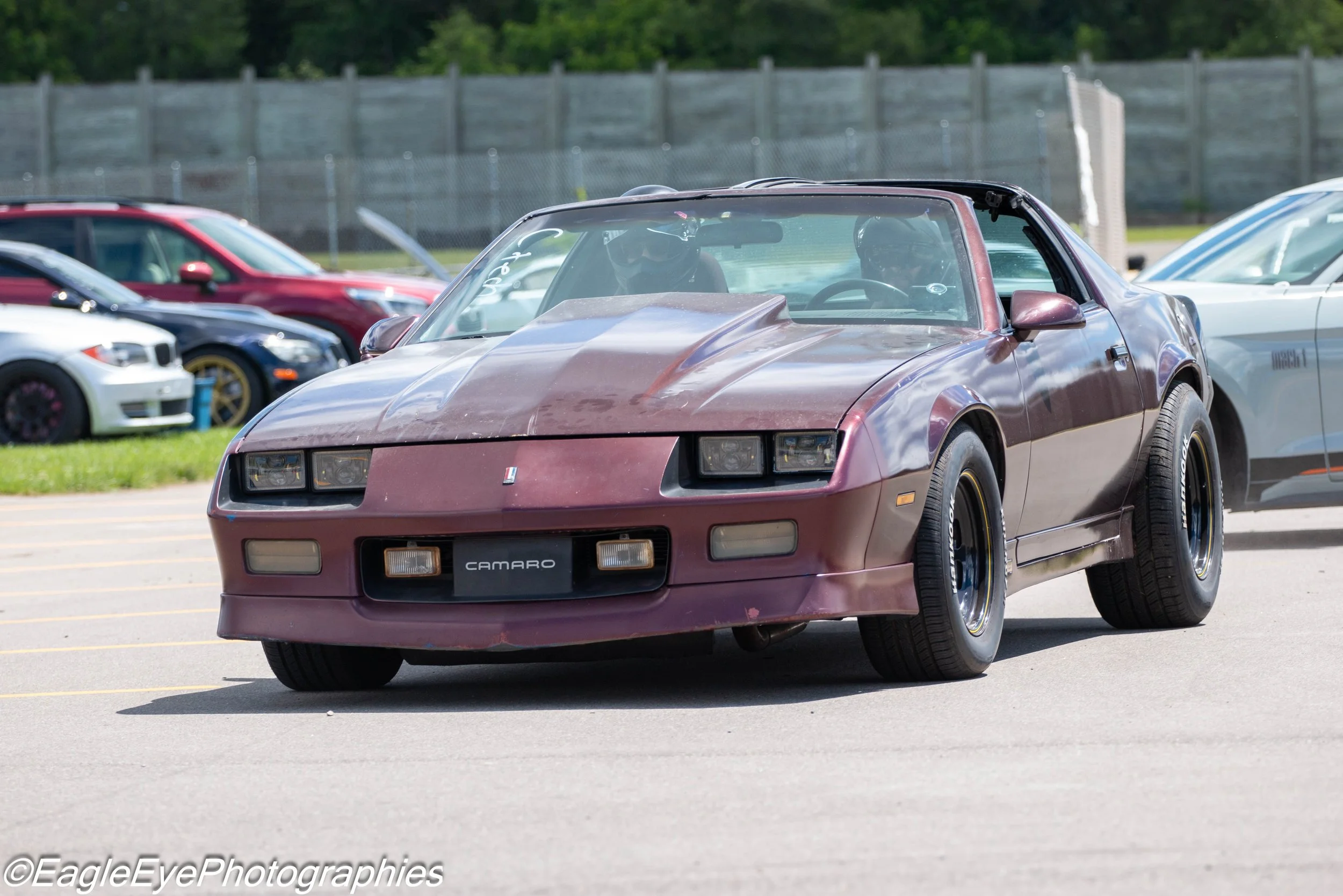 A vintage maroon Chevrolet Camaro convertible driving in a parking lot, surrounded by other cars, with a person in the driver's seat wearing a helmet.