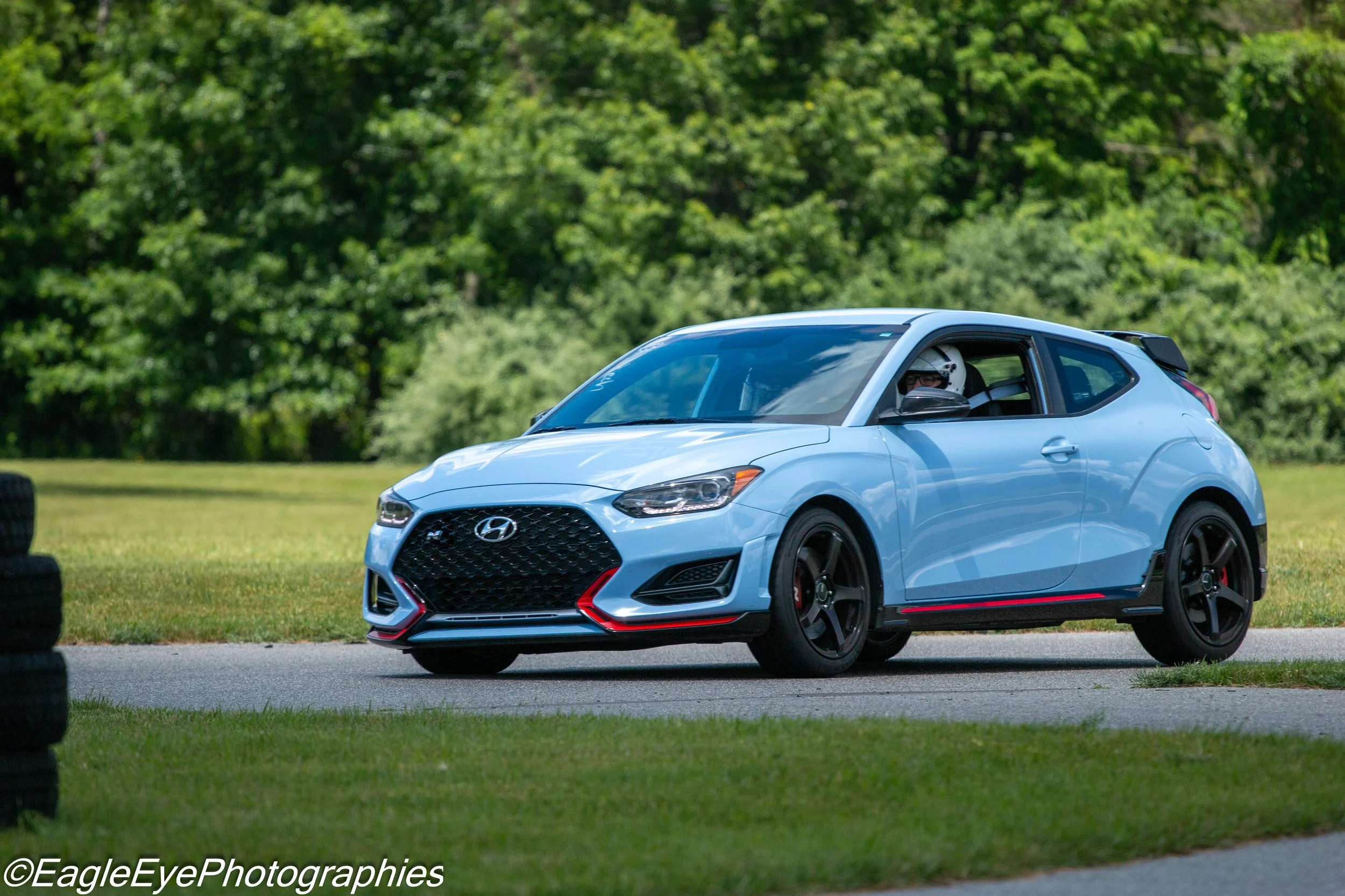 A light blue Hyundai car with a driver wearing a helmet, parked on a grassy area with trees in the background.