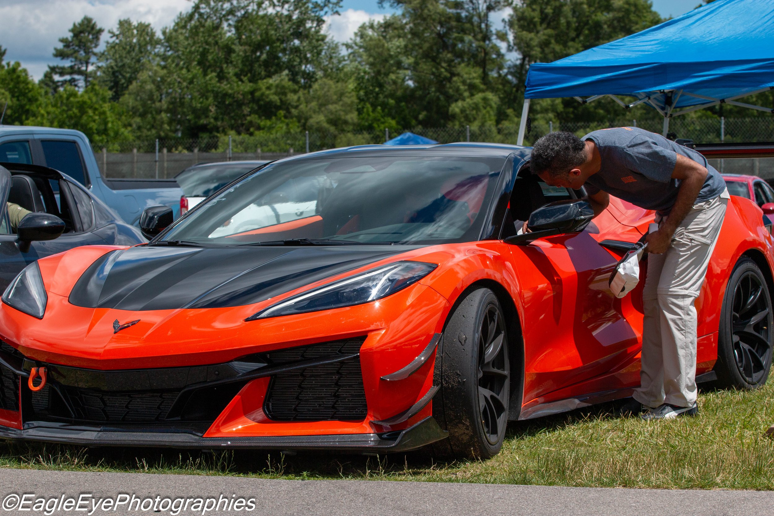 A man inspecting an orange and black sports car at an outdoor car show with tents and a fence in the background.