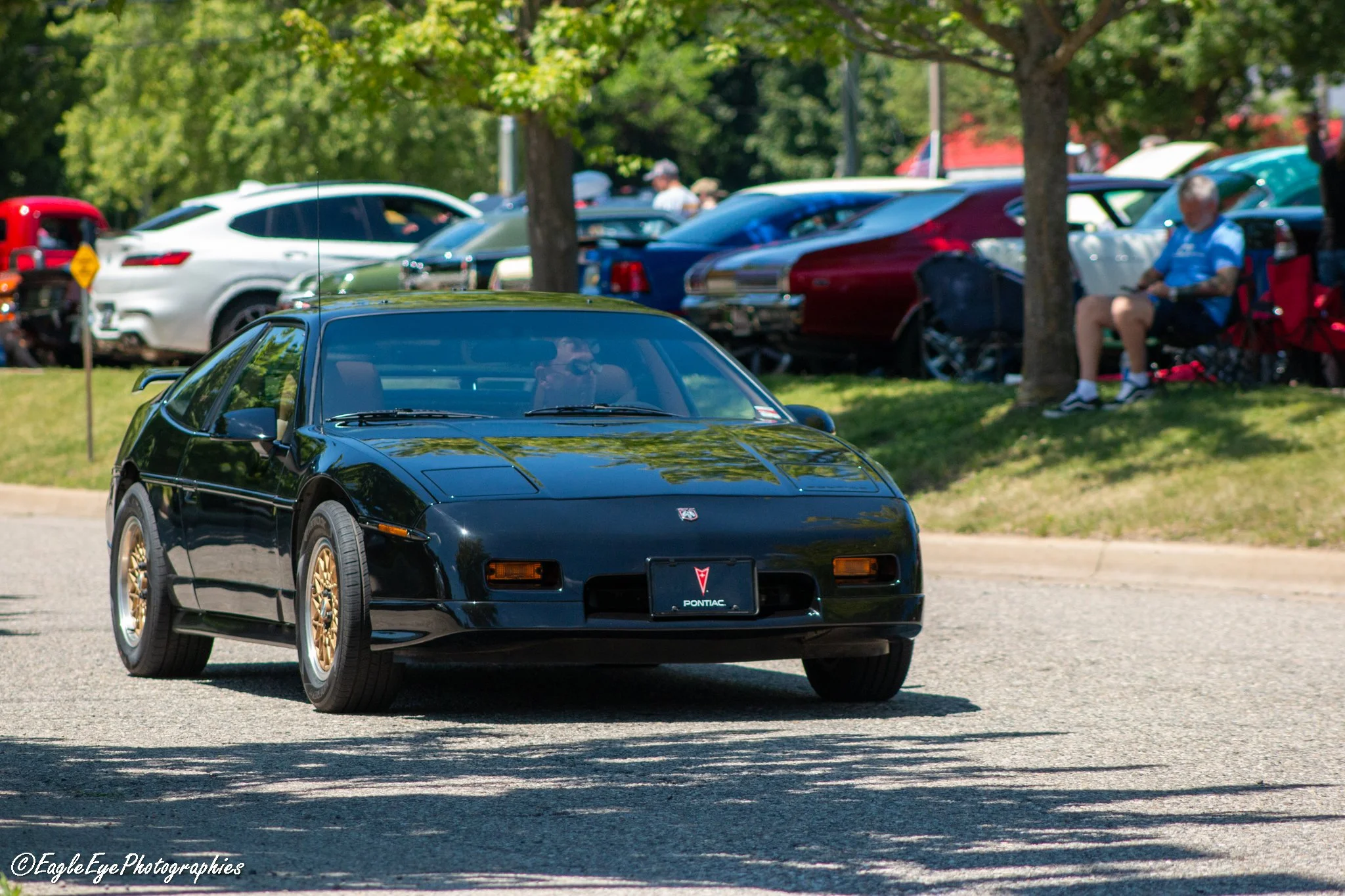 A black Pontiac sports car driving on a paved road during a car show, with multiple parked cars and spectators under trees in the background.
