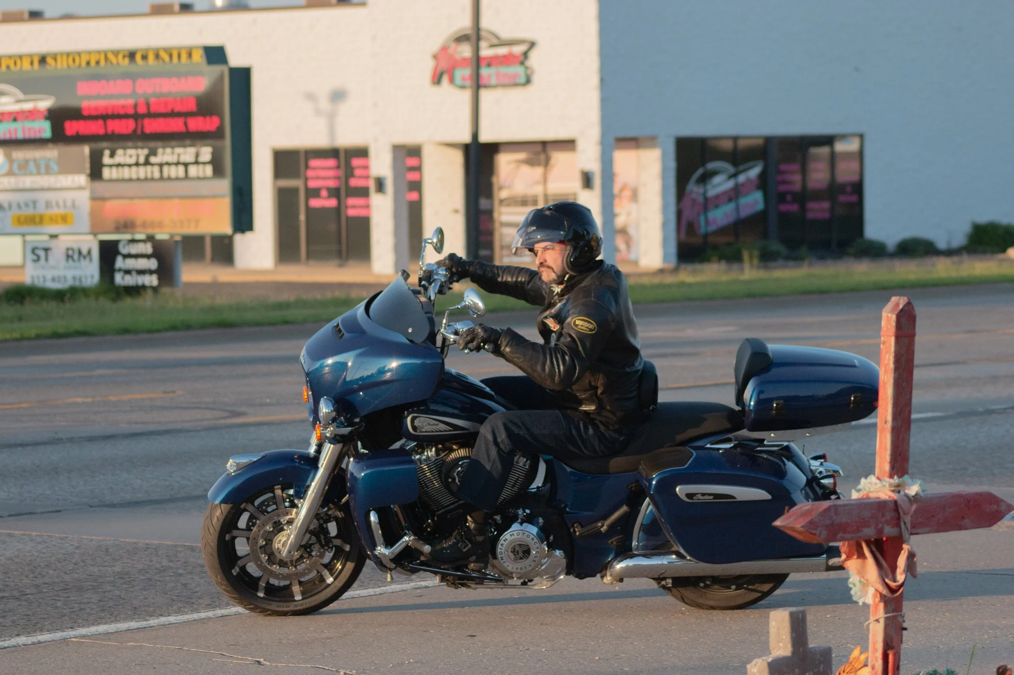 A man wearing a black leather jacket, black helmet, and glasses rides a blue motorcycle with a sidecar on a city street. The background shows a building with signs for retail stores and a parking lot.