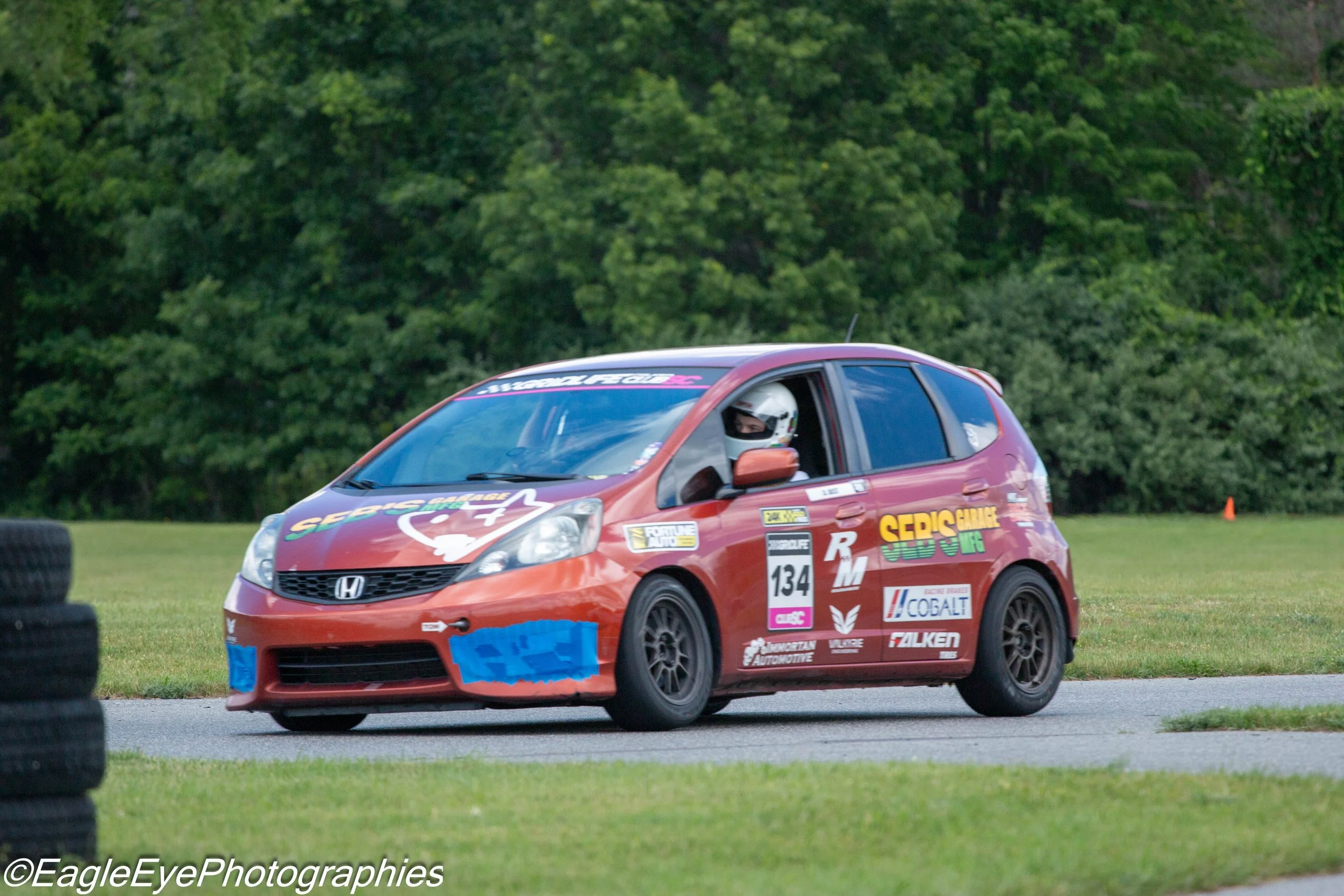 Red race car with driver wearing helmet on a grassy track, surrounded by tires, and a green tree backdrop.