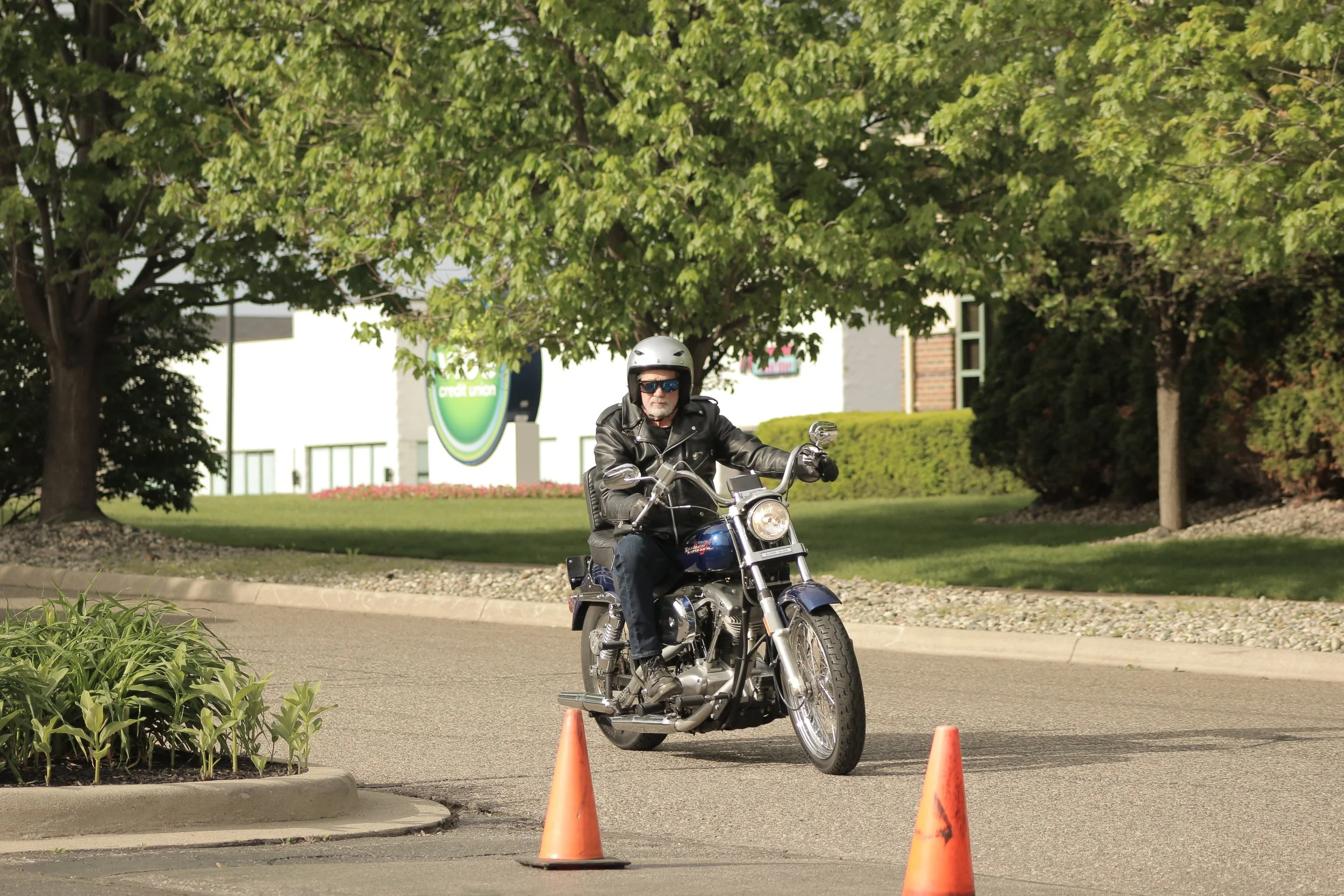 A man wearing sunglasses, a black leather jacket, and a helmet rides a motorcycle through a parking lot separated by orange traffic cones, with green trees and buildings in the background.