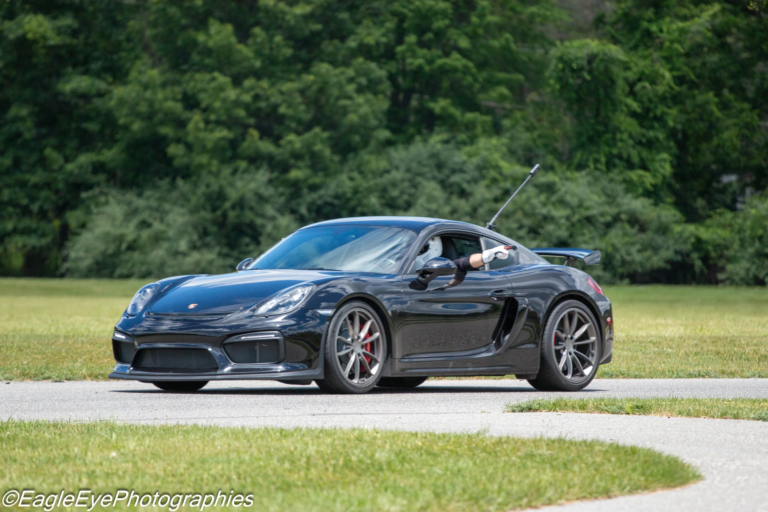 A sleek black sports car driven by a person wearing a helmet and gloves, racing on a track with a forested background.