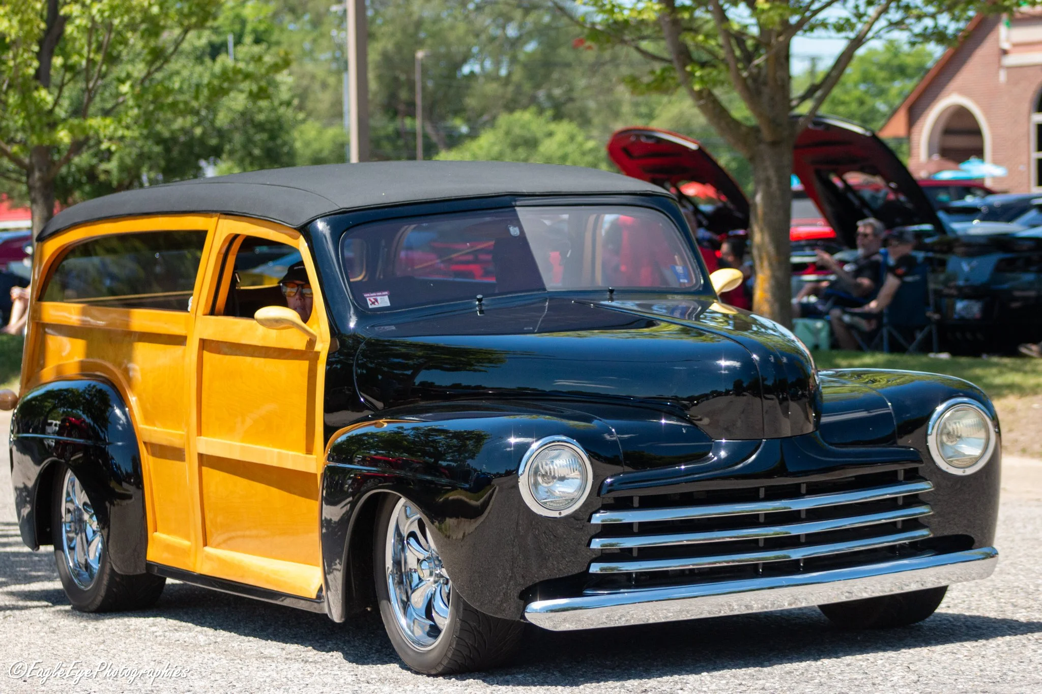 A vintage black and yellow car at an outdoor car show with trees and other cars with open hoods in the background.