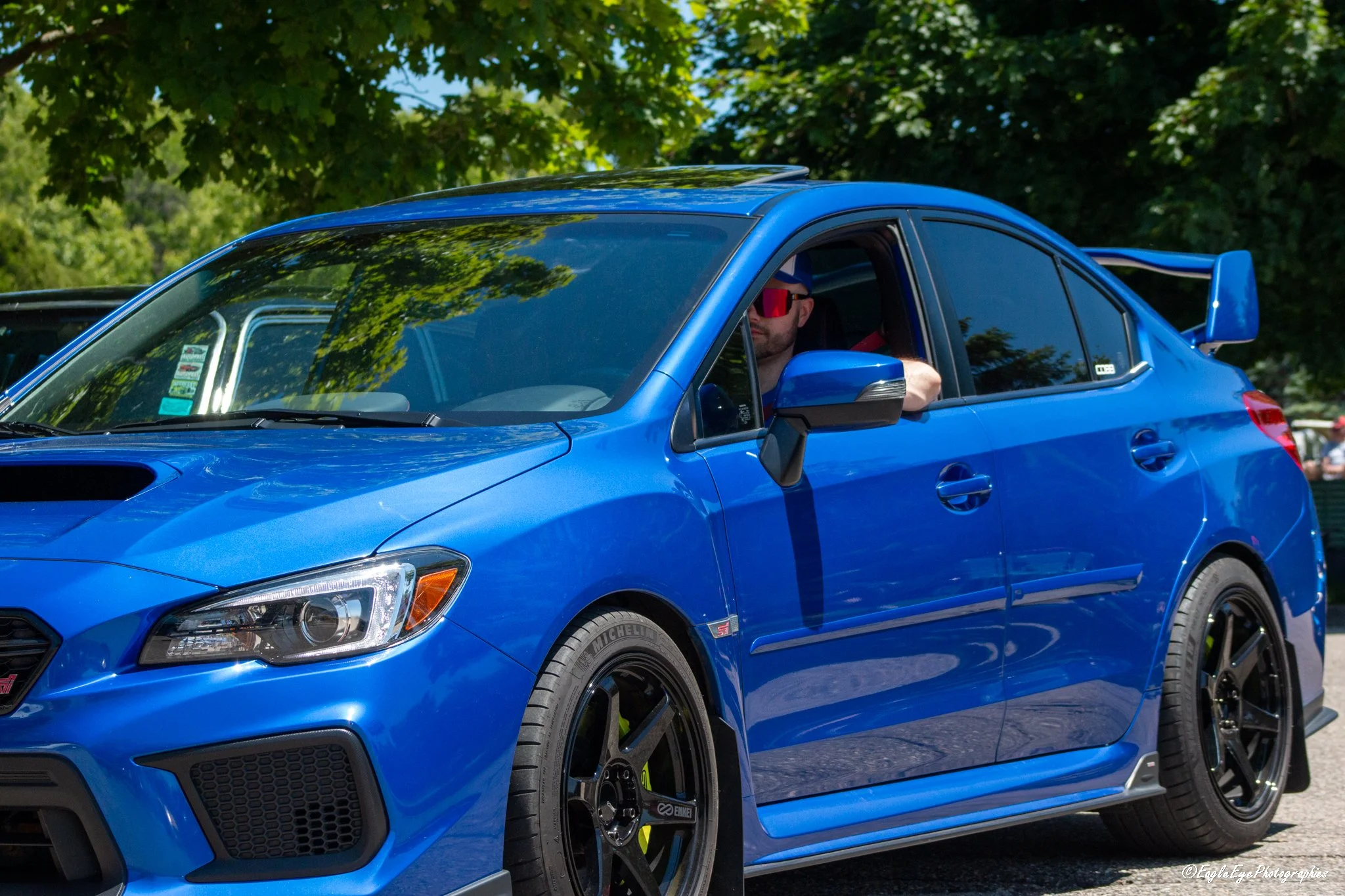 A blue car parked outdoors beneath green trees with sunlight filtering through the leaves, featuring a driver wearing sunglasses.
