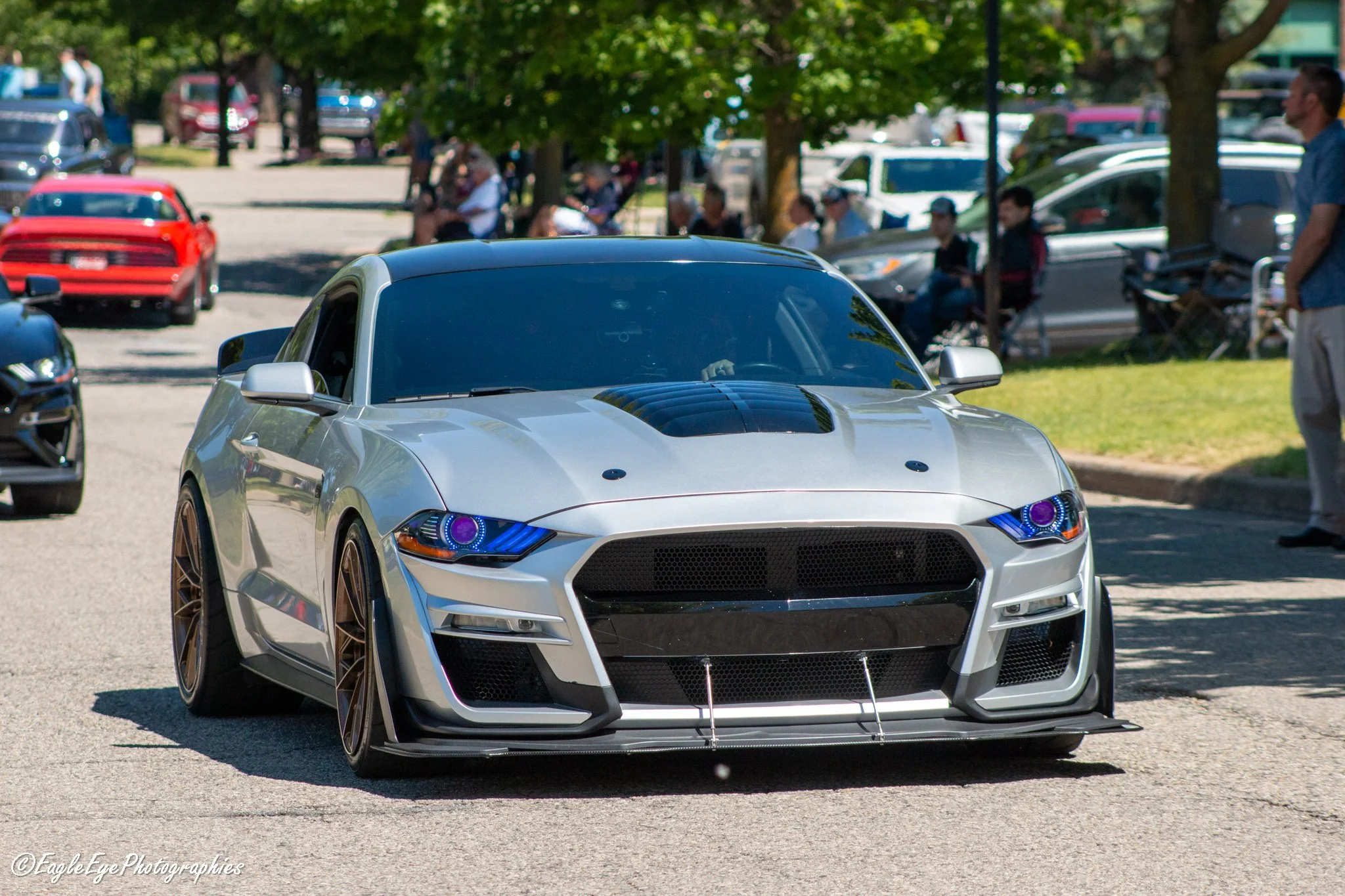 A silver sports car with custom wheels and headlights, parked outdoors at a car show on a sunny day, with people and other vehicles in the background.