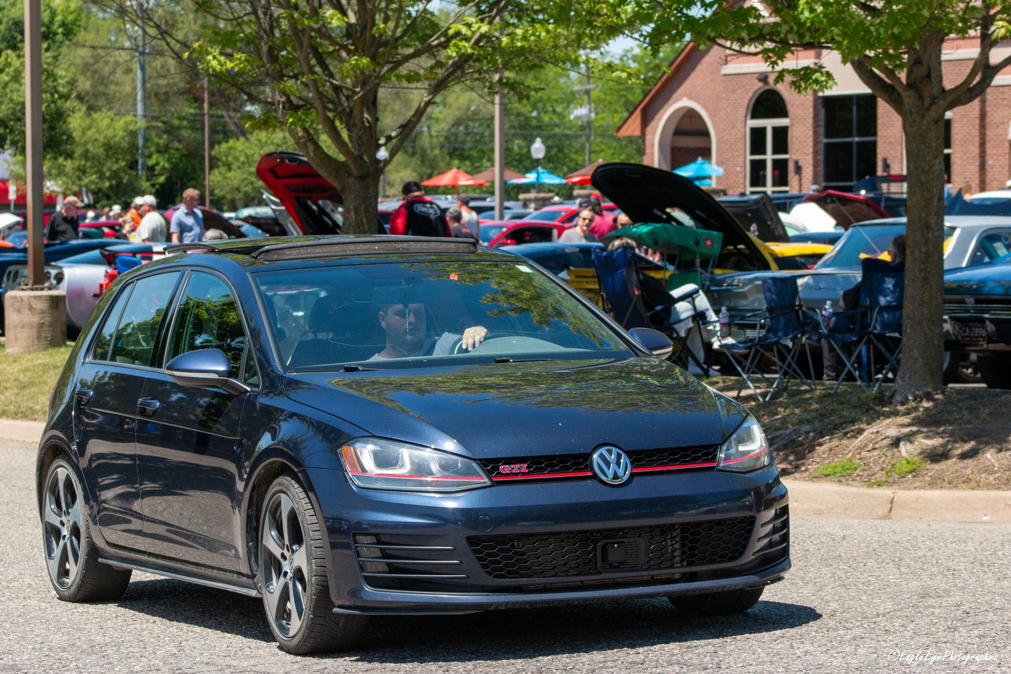 A black Volkswagen Golf GTI parked in front of a crowd at an outdoor event with cars and people inspecting them, surrounded by trees and a brick building.