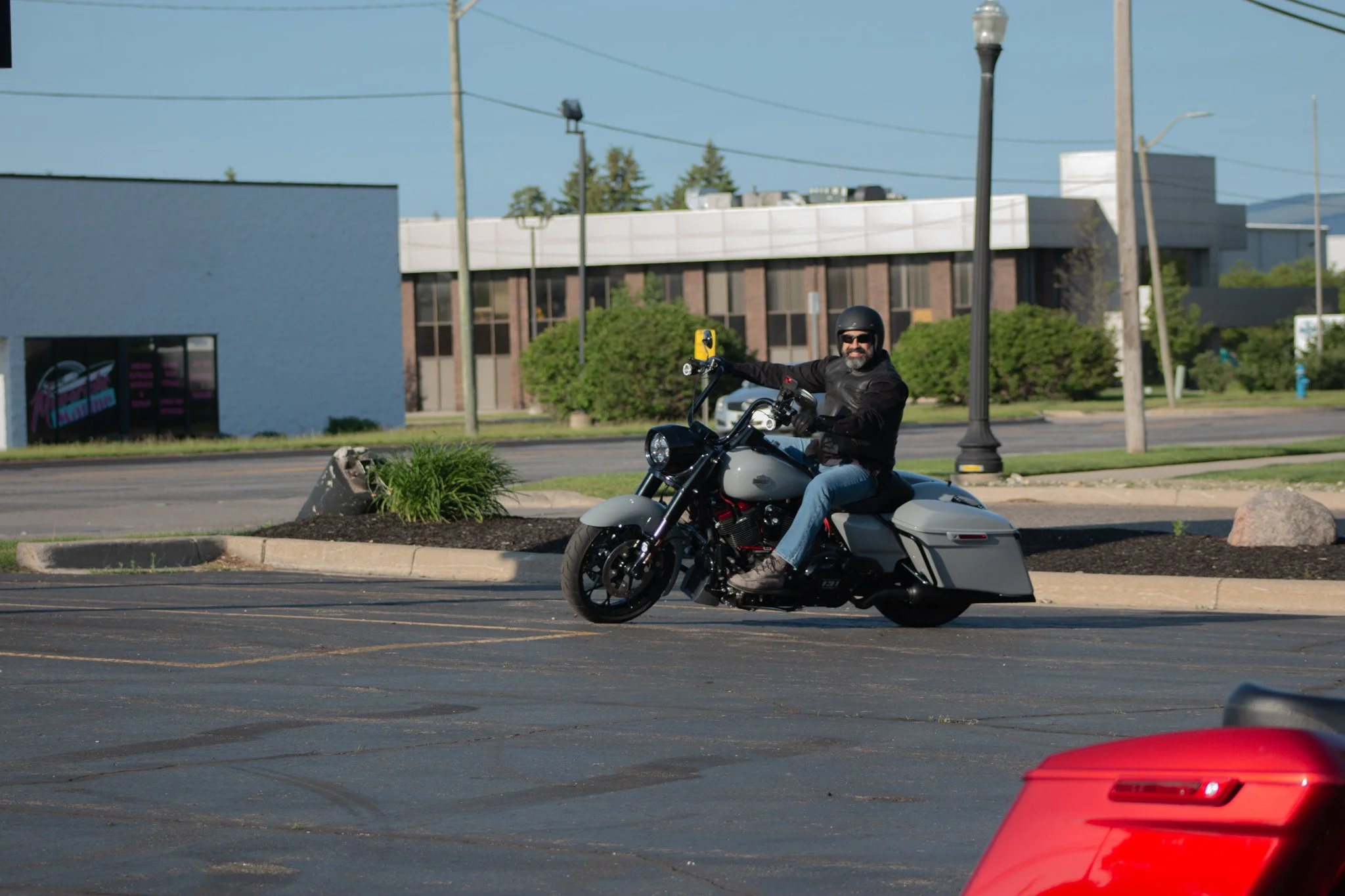 A man with a beard, wearing sunglasses, a black helmet, a black leather jacket, and jeans, riding a gray motorcycle in a parking lot with a cityscape backdrop.