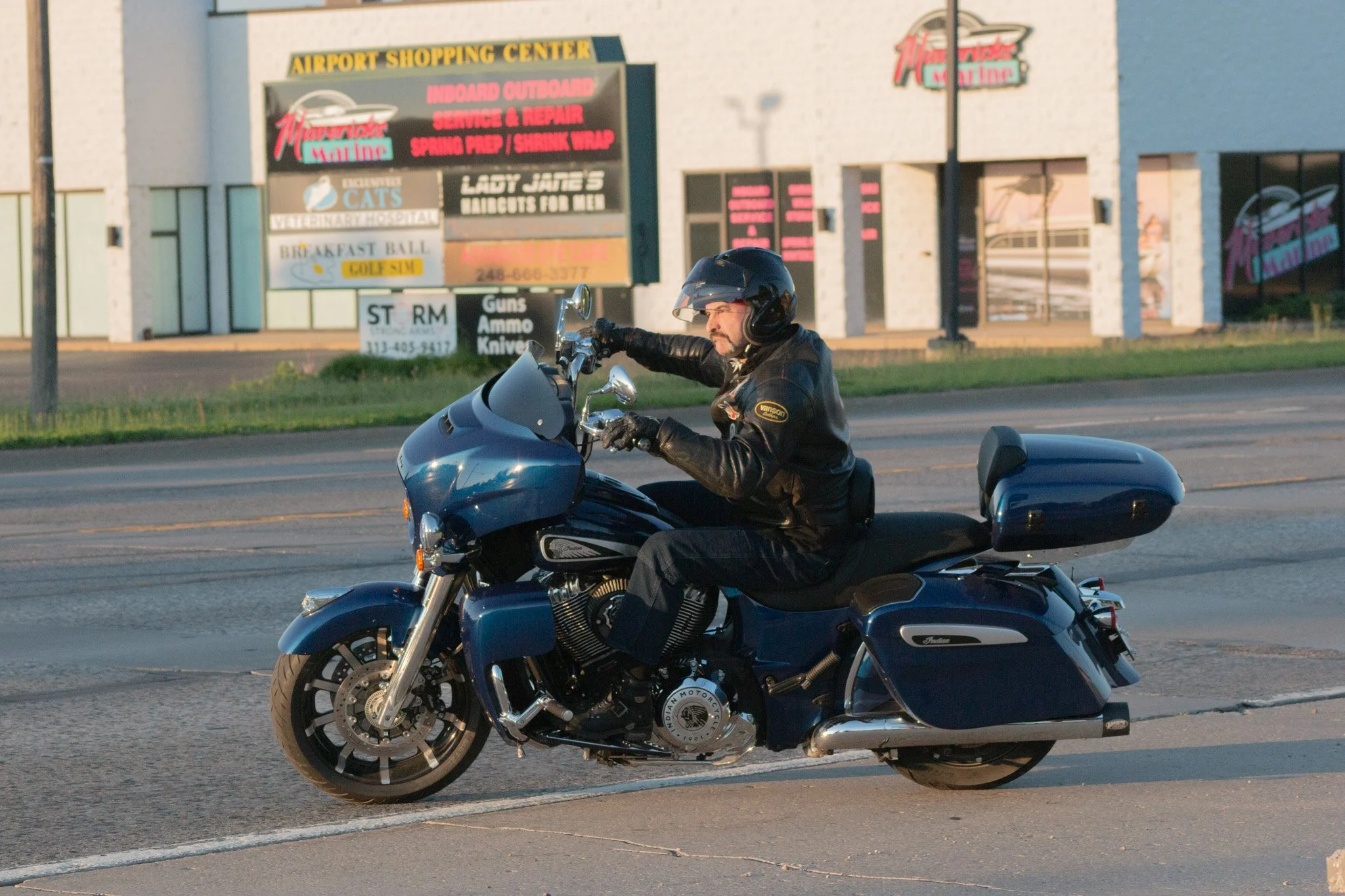 A man riding a black motorcycle on the street in front of a shopping center with a signboard advertising various services and shops.
